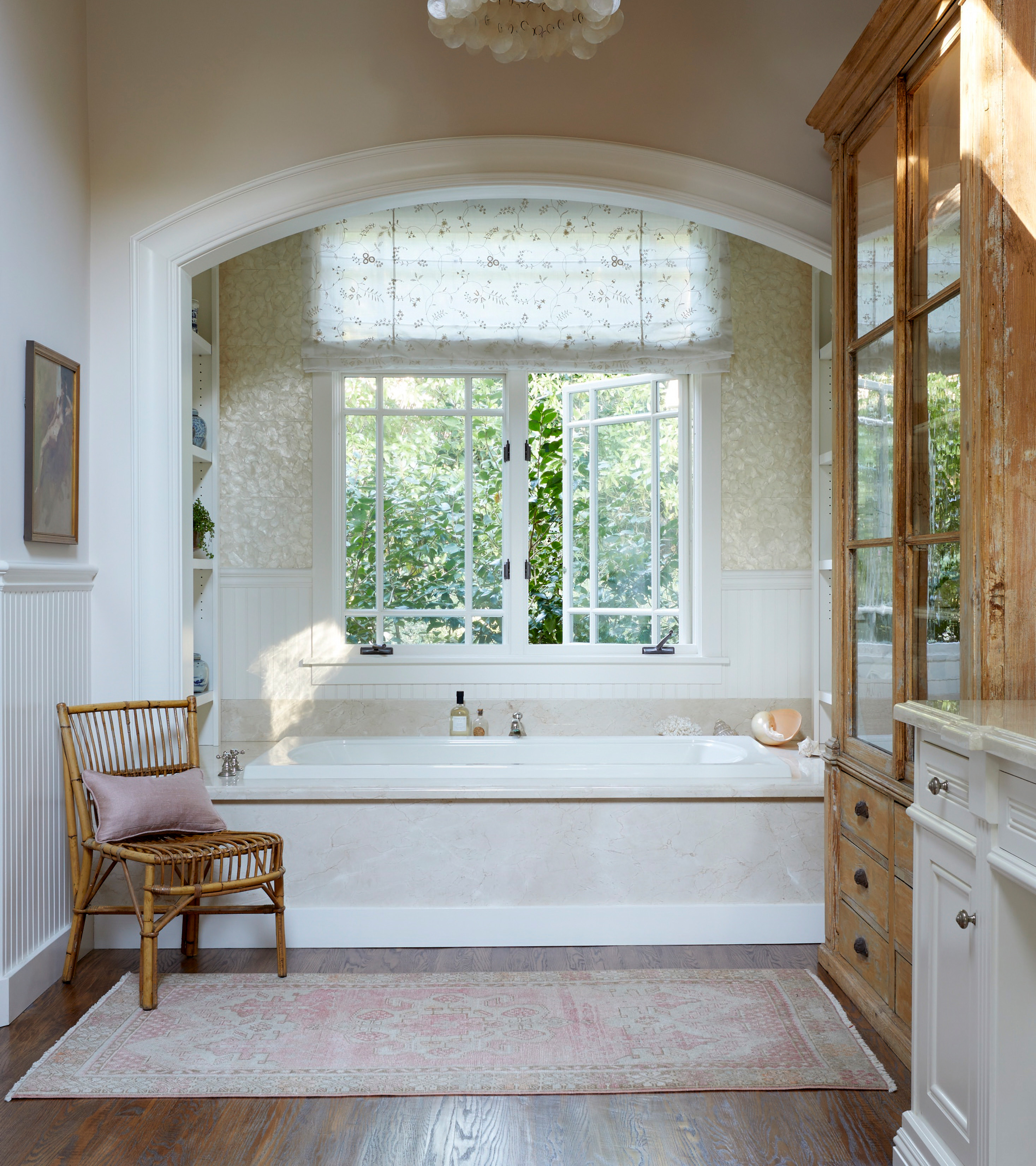 a bathroom with a built in bath nook in light marble, a large window with white floral blinds, a rattan chair next to the bath, a pale red rug on dark wood floor and a tall wooden display cabinet