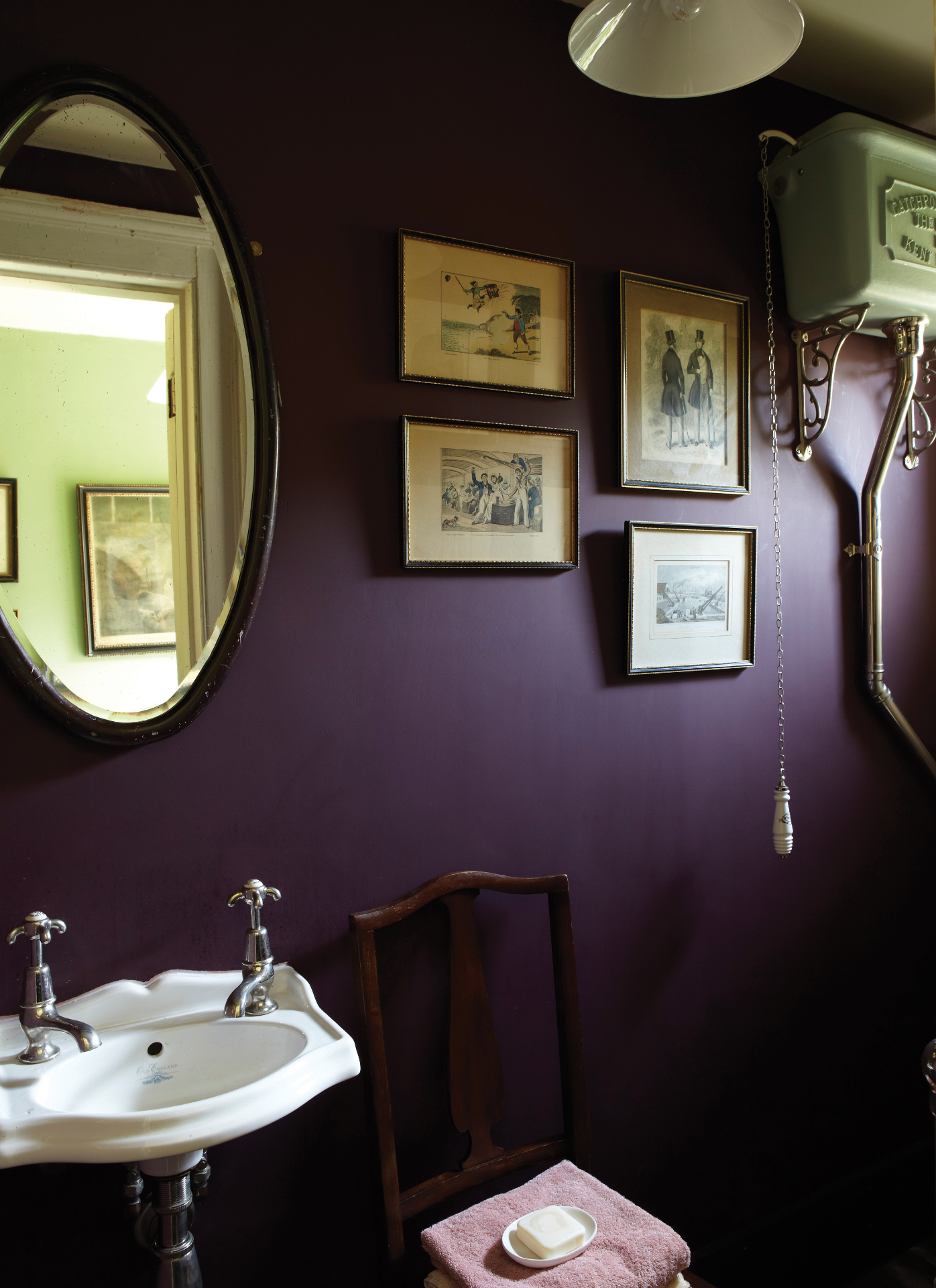 a bathroom with dark purple walls, a collection of vintage illustrations in frame and a high level sisterne with a pull lever