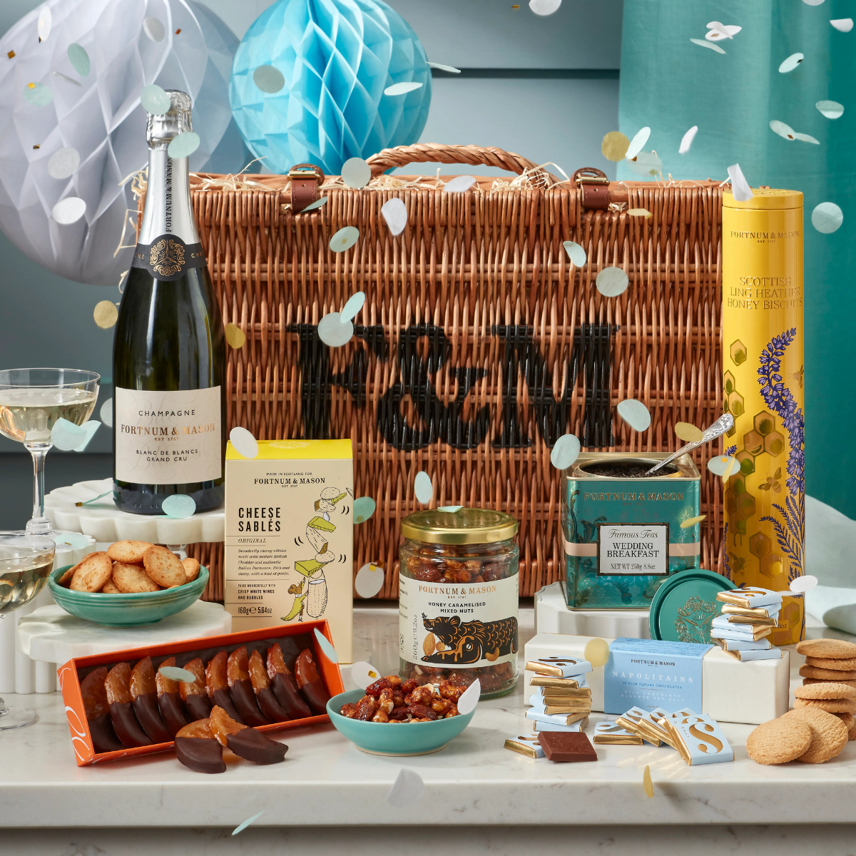 A spread of biscuits, nuts, chocolate, tea and champagne in front of a wicker fortnum and mason hamper
