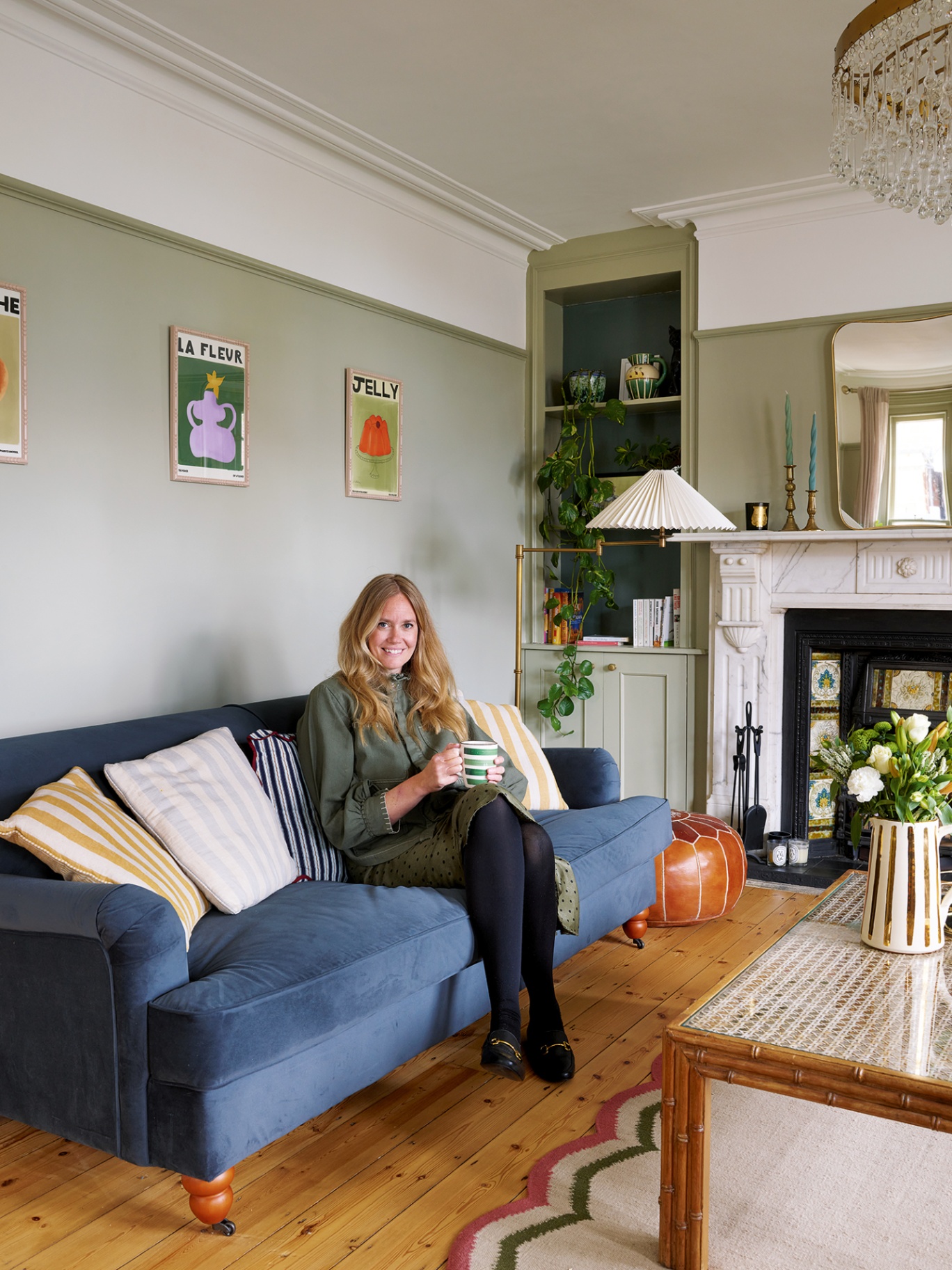 A woman sat holding a cup of tea on a blue velvet sofa, leaning on a selection of striped, colourful cushions. Behind a tall brass floor lamp topped with a pleated shade stands in front of built-in shelving. The walls are painted in pale green.