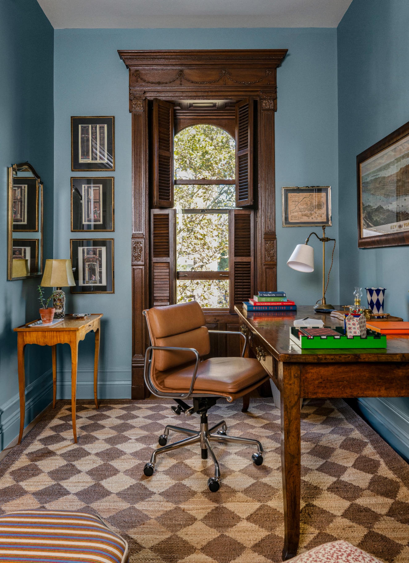 Blue-painted study with dark wood carved window shutters, a vintage wooden desk, modern cognac leather swivel desk chair, and a beige and brown checkerboard rug