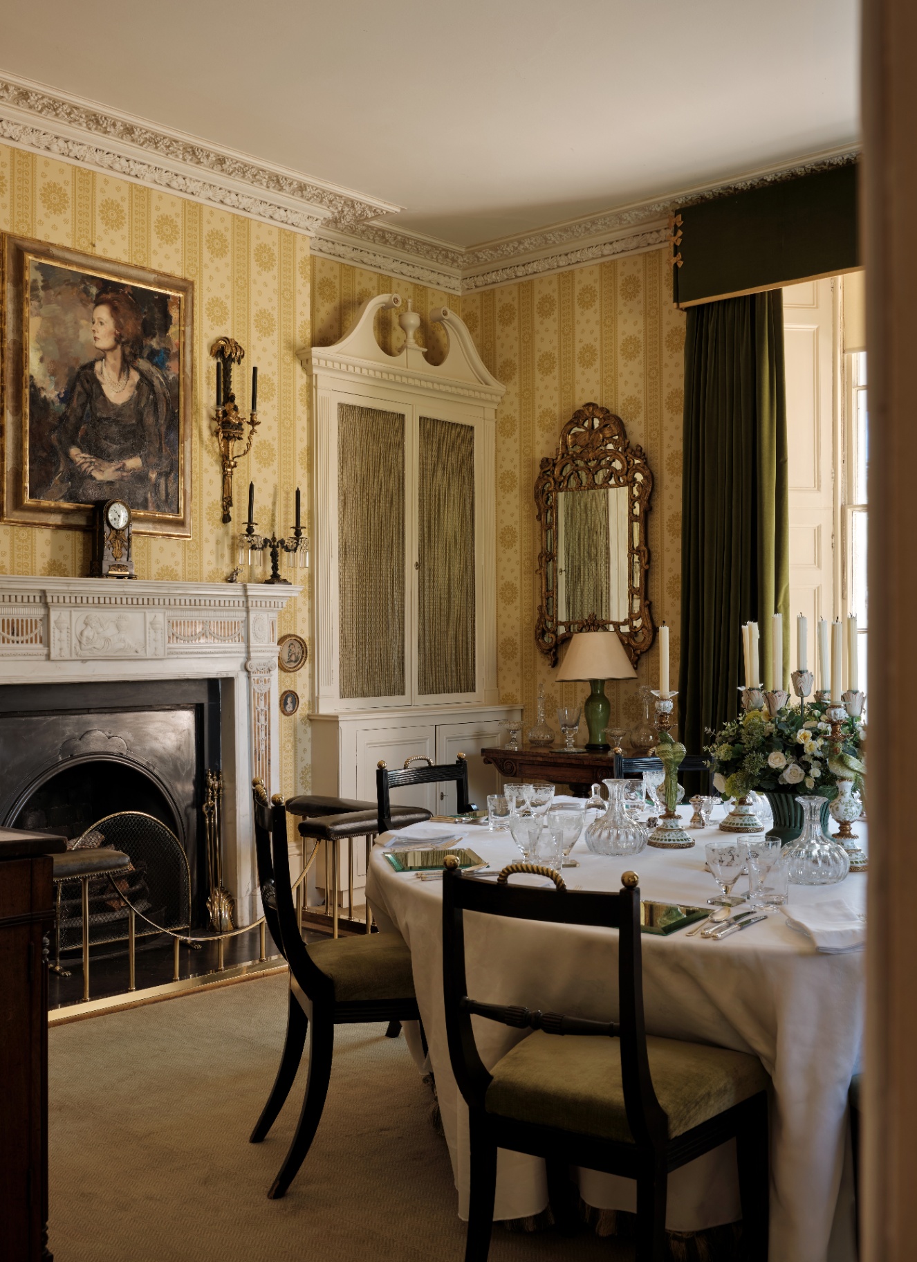 Traditional dining room with yellow patterned wallpaper, dark green drapery, and a formal table setting illuminated by table lamps and tall candles