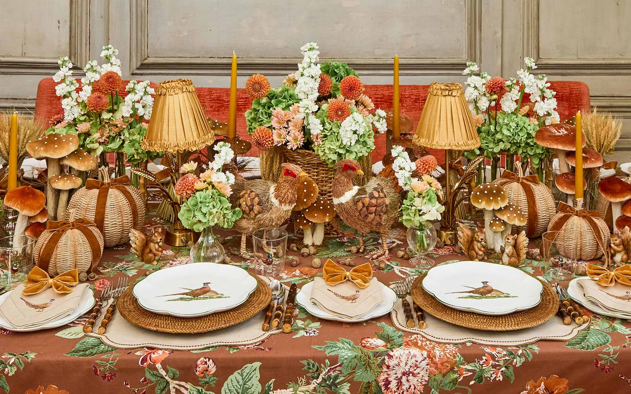 an autumn table with a brown floral tablecloth, white plates with pheasant illustrations, rattan charger plates, rattan vases of flowers and pheasant, pumpkin and mushroom ornaments