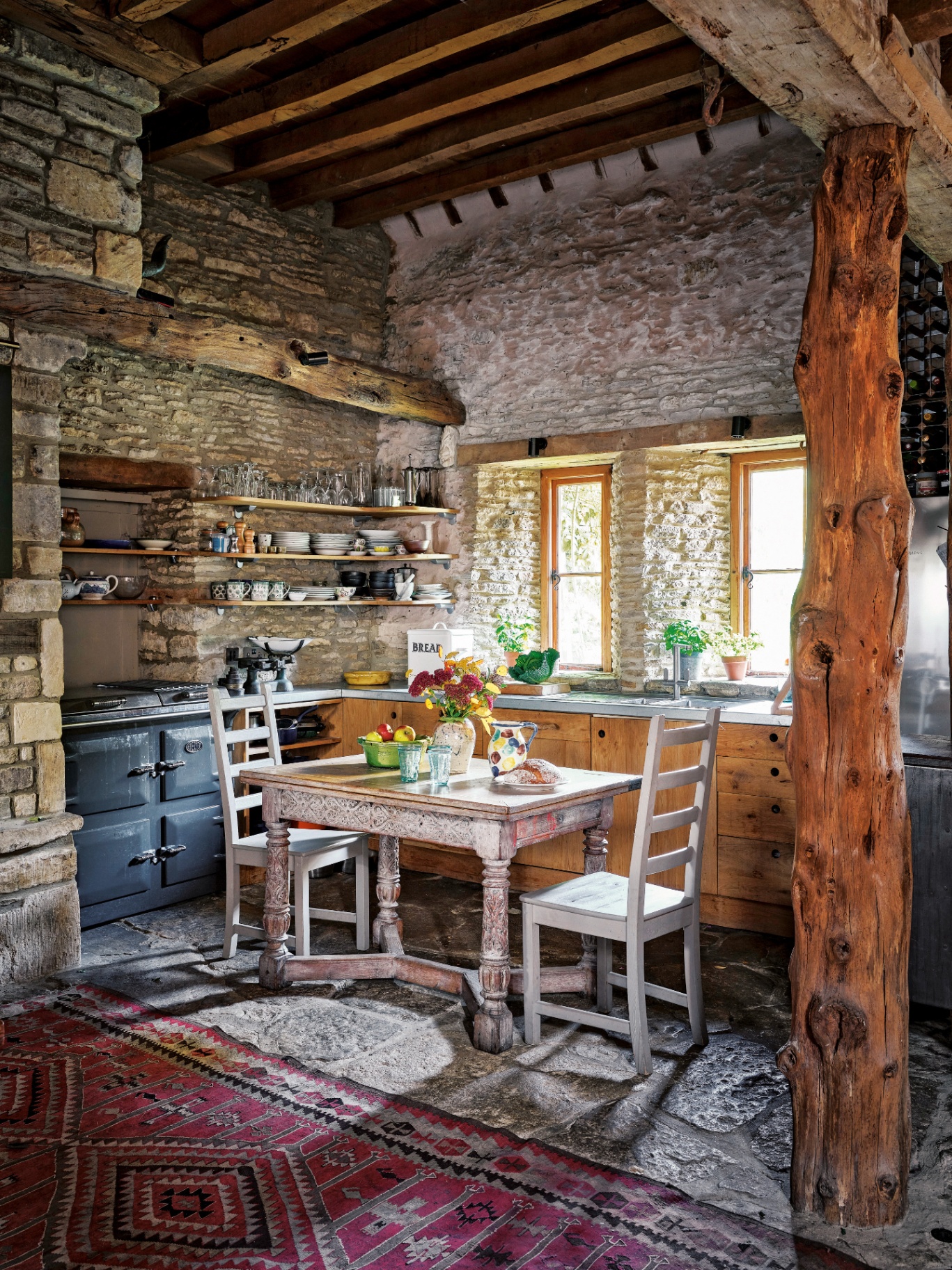 a kitchen with exposed brick and old wooden beams, a red patterned rug, a small dining table and chairs, a range cooker and open shelves