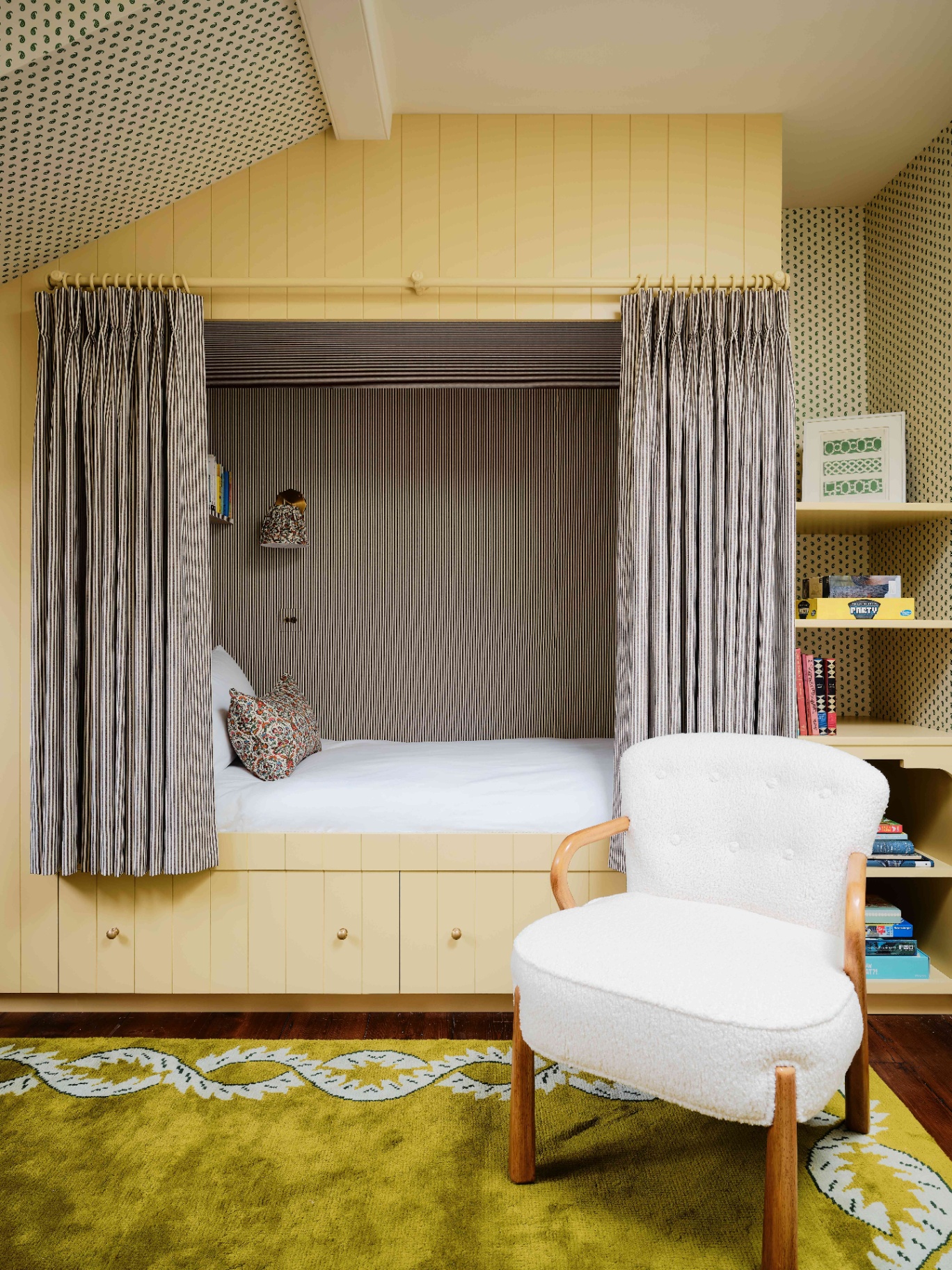 a children's bedroom with an alcove bed painted in lemon yellow, black and white striped curtains covering the bed alcove, built in open shelving to the right of the bed, a chartreuse patterned rug and a boucle accent chair