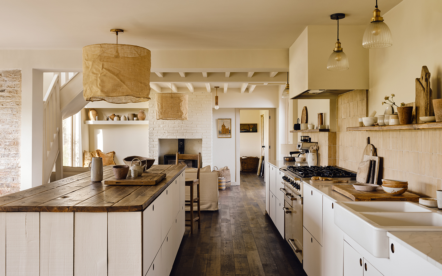 An open plan kitchen with an wood-topped island, white cabinets, cooker and sink in the foreground. In the background, white linen sofas are arranged opposite a woodburner.