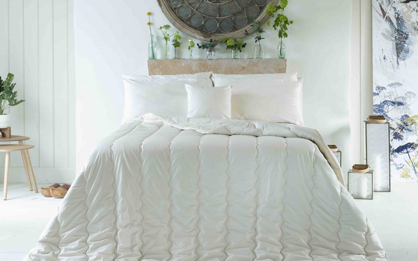 a white bedroom with a double bed with a duvet on top, two lanterns next to it and a large print above the bed with a shelf full of bud vases and greenery