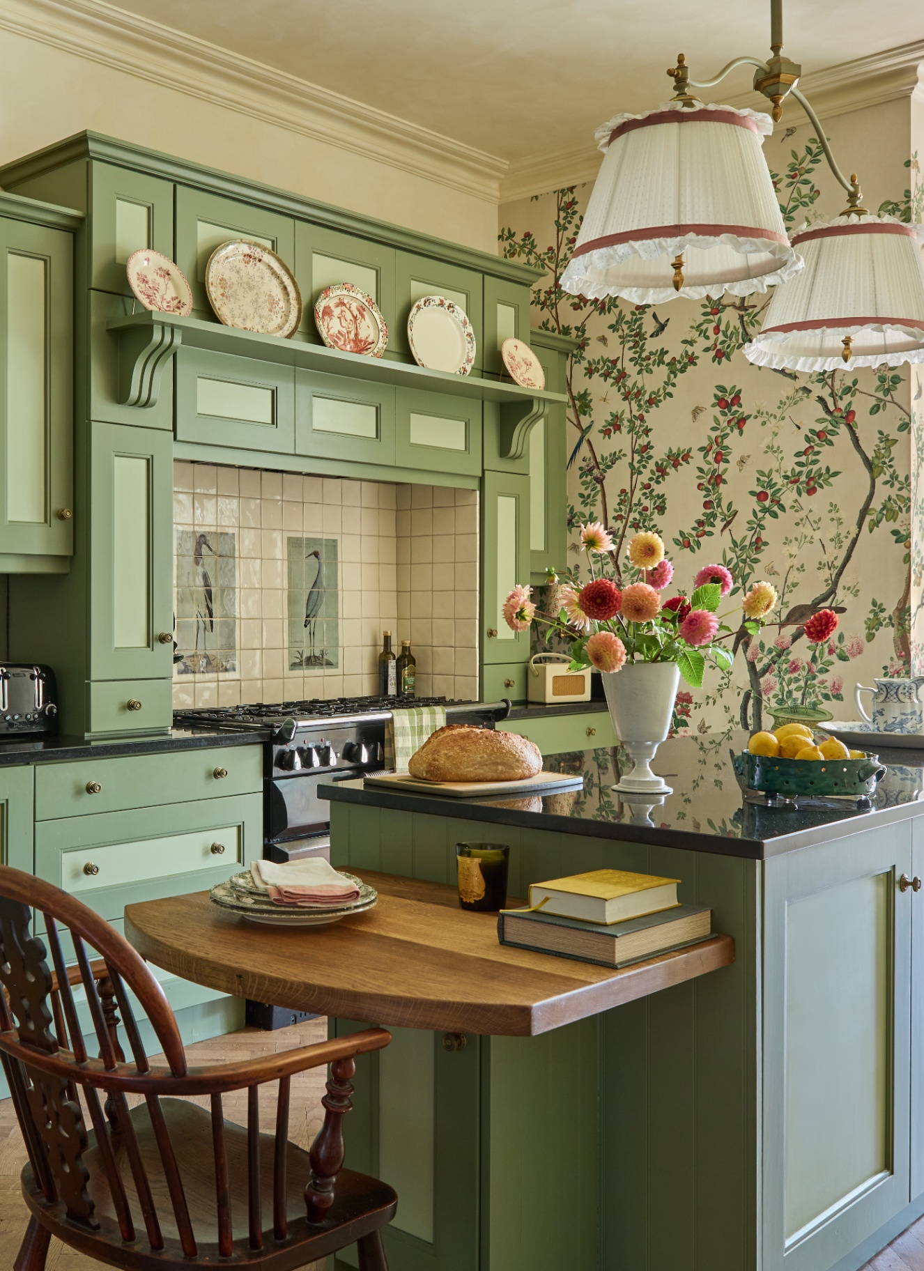 a kitchen with floral wallpaper, a sage green island with a drop leaf table and a spindle chair and arange cooker with a bird tiled splashback