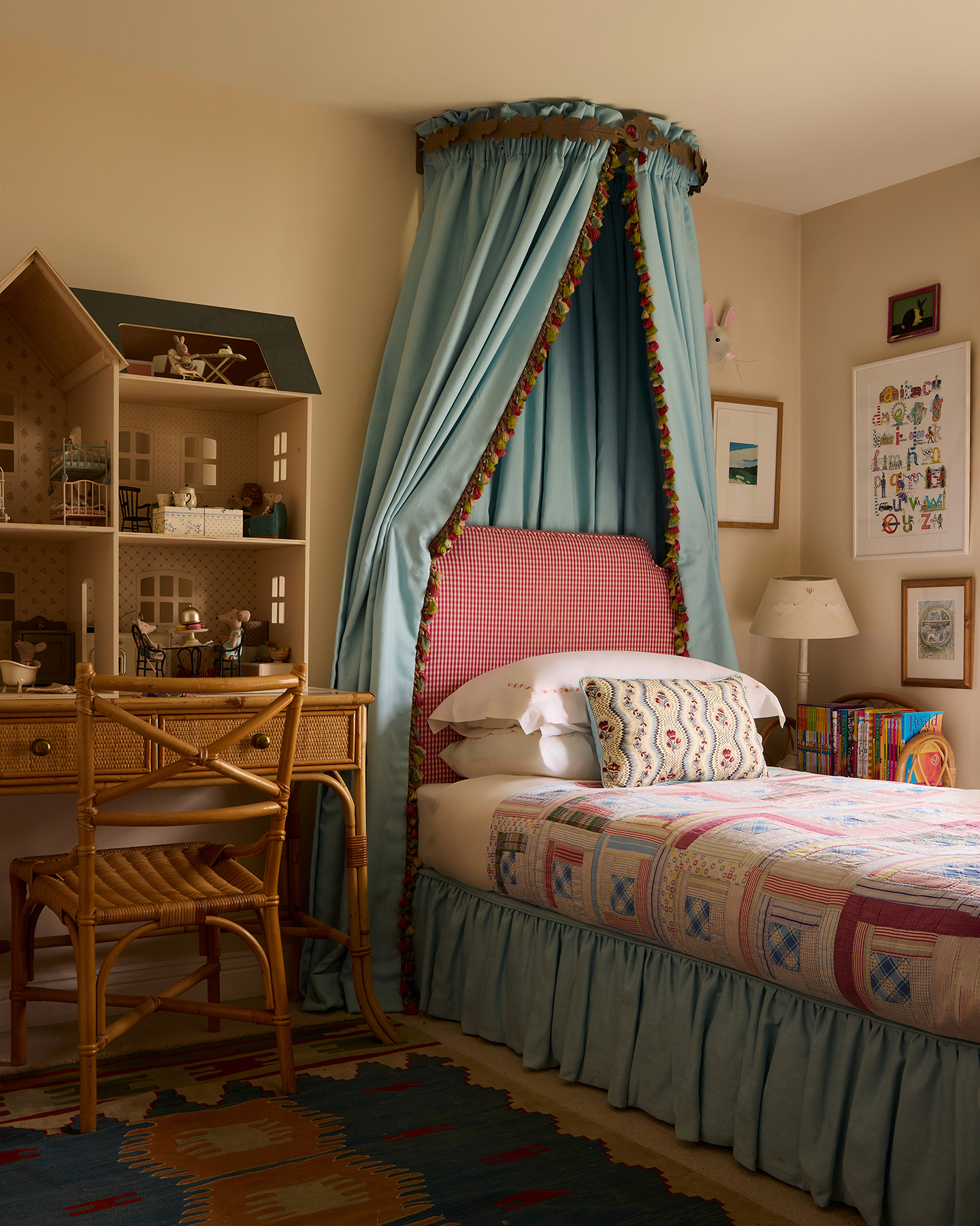 A children’s bedroom with a single bed topped with a tasselled canopy and with a matching bed skirt. A desk and chair sit to the side with a large dolls house on top