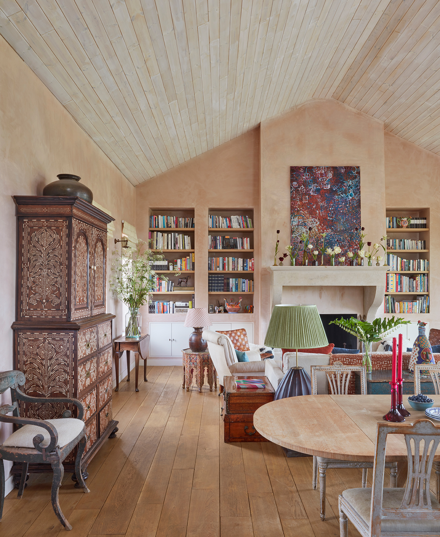 A high ceilinged-living room with timber-clad ceilings and raw plaster walls. In the foreground is the end of a wood dining table with chairs and a decorative cabinet, and in the background a colourful seating area surrounds a fireplace