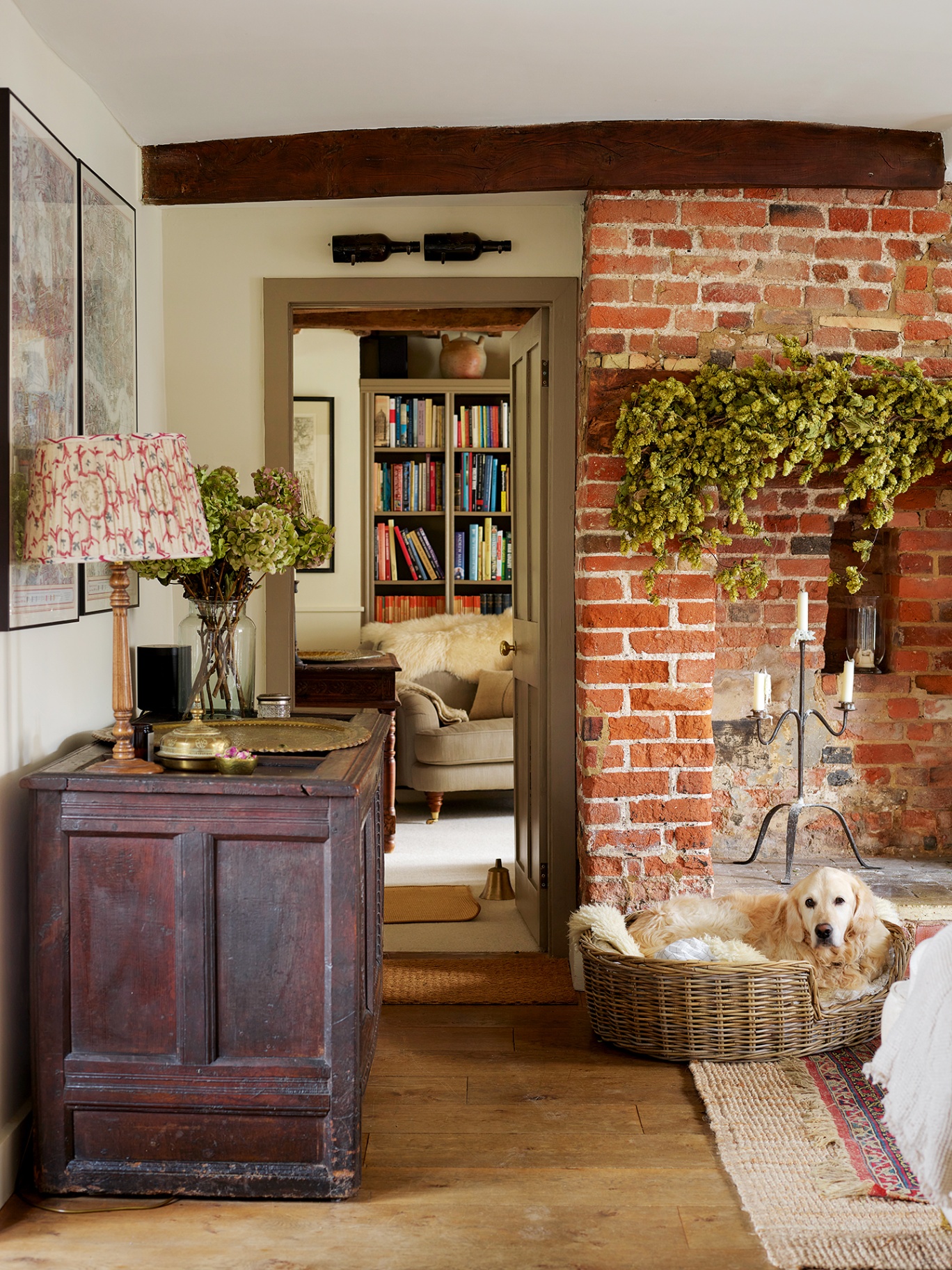 A living room with large brick hearth decorated with hops and a golden retriever in a dog basket in front. A large antique chest lines the wall on the left, topped with a lamp and vase with flowers. An open doorway looks onto an armchair and bookshelves