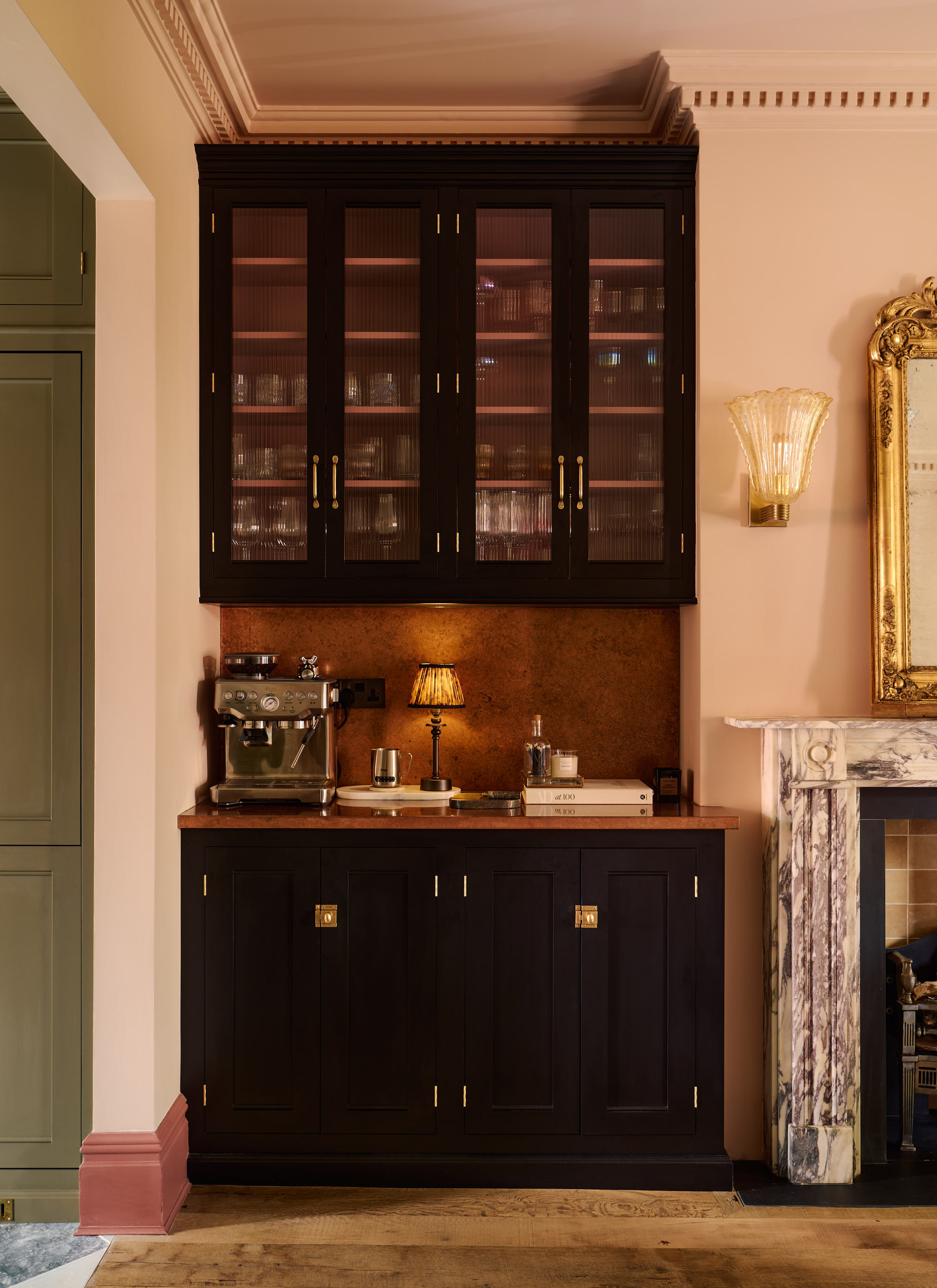 a room with pale pink walls, wooden flooring, a white and grey marble fireplace with a gold mirror above it and a black built in cupboard with a silver coffee machine, a book and a pleated lamp on top and a black floating cupboard above it