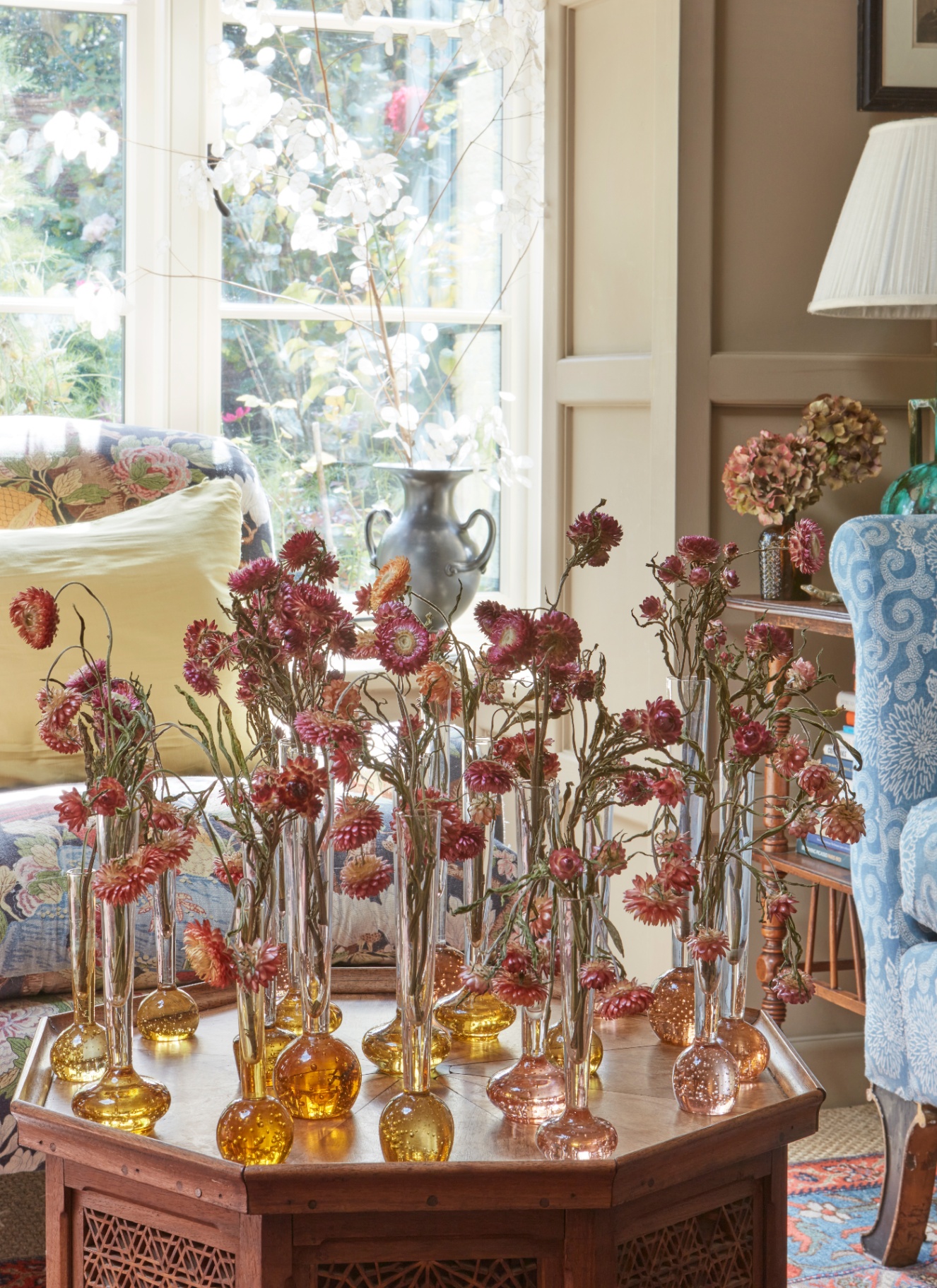 A collection of small, tinted glass bud vases filled with dried, rust-colored strawflowers, tightly clustered on an octagonal wooden coffee table