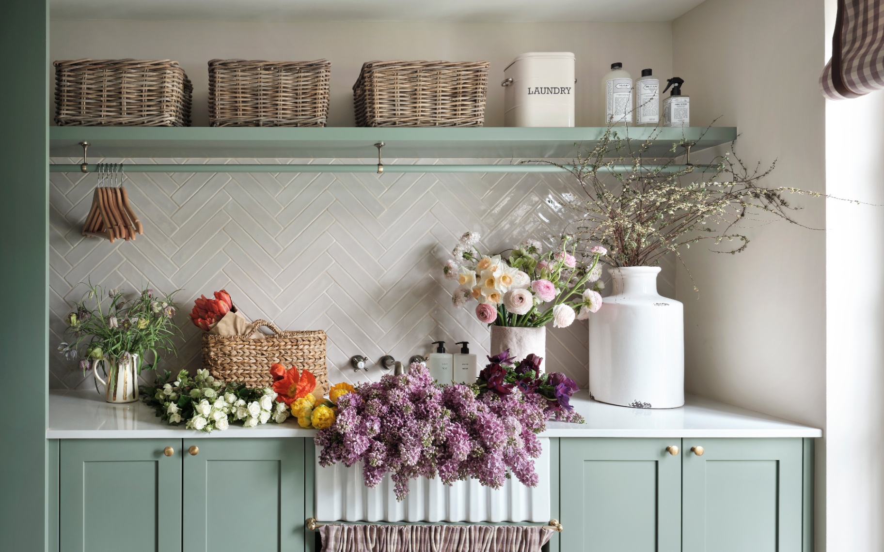 a utility room with sage green cabinets, a belfast sink filled with fresh flowers and a sage green open shelf with wicker baskets