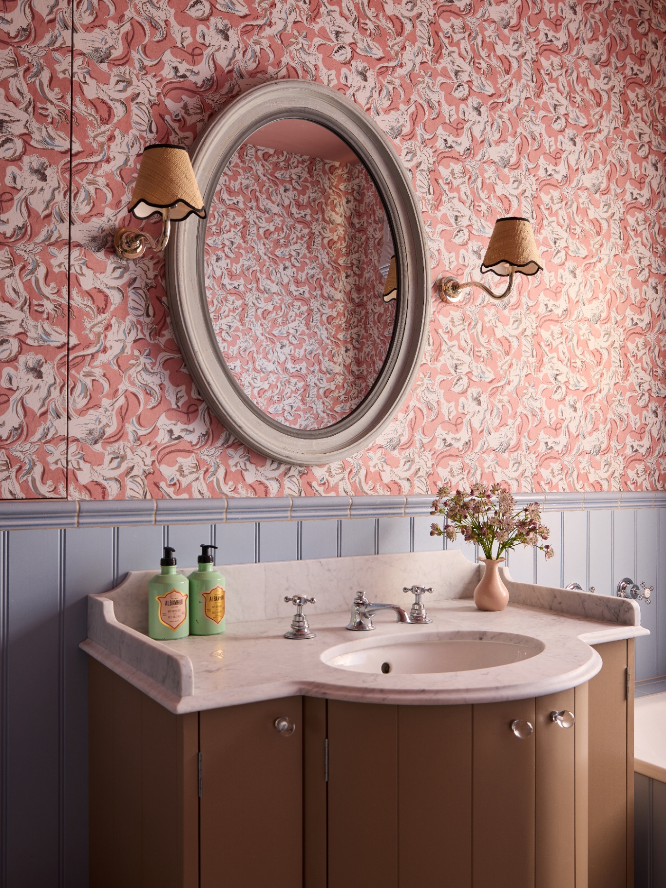 a custom made vanity pictured for a bathroom with a large wood oval mirror hanging above it on a pink patterned wallpaper 