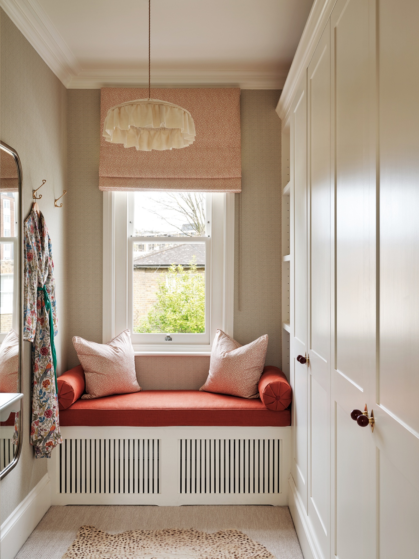 A narrow dressing room with built-in wardrobes running along the right side, a window seat with orange seat cushion and bolster cushions. Above is a window fitted with a roman blind and a frilly pendant lamp. A floral dress hangs next to a mirror