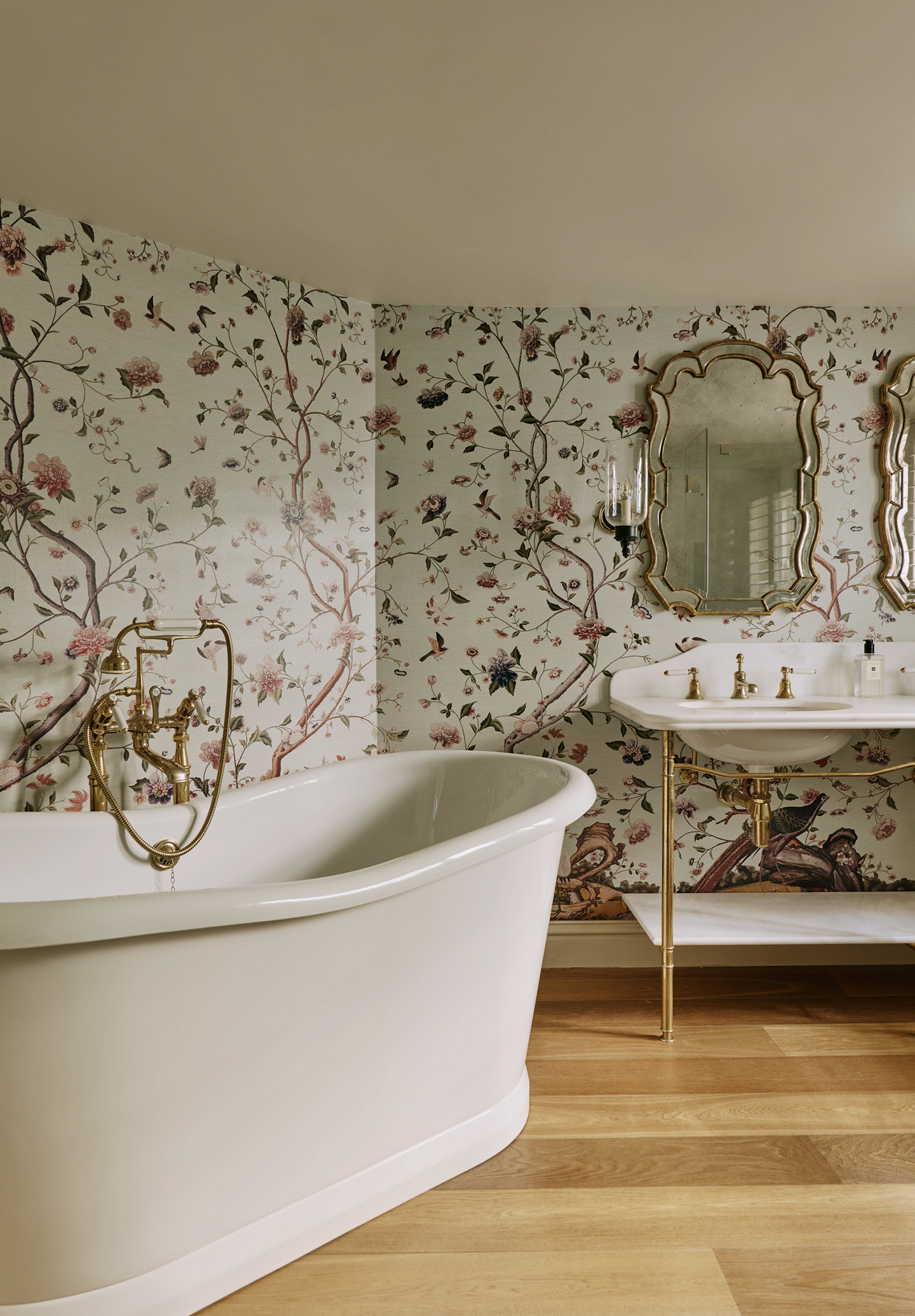 The corner of a bathroom with a freestanding bath tub and traditional wall-fixed double basin, with two ornate mirrors and a wall light above. A botanical mural wallpaper covers the walls. 