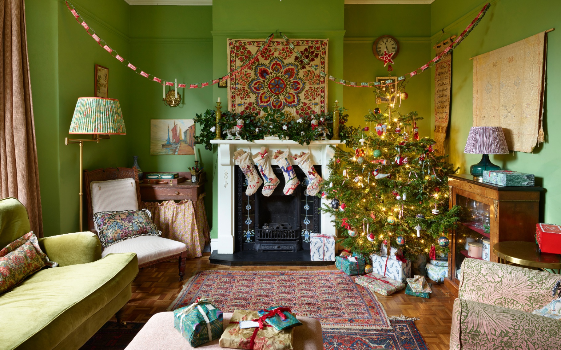 An apple-green sitting room featuring personalized embroidered stockings on the mantelpiece, paper garlands, and a lit Christmas tree surrounded by wrapped gifts