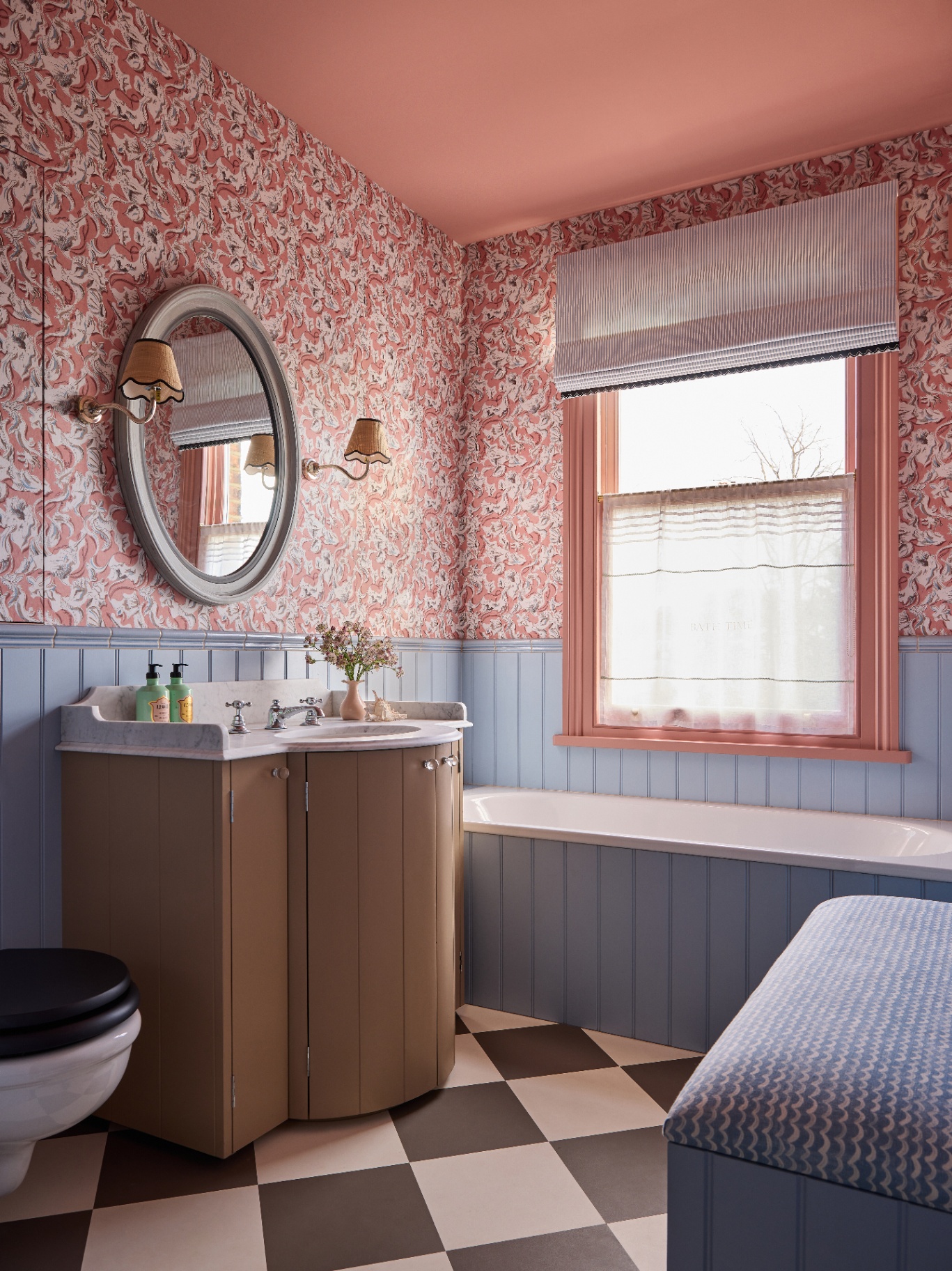 a pink wallpapered bathroom and painted ceiling pictured with checkerboard flooring, a small vanity with a mirror is at the centre