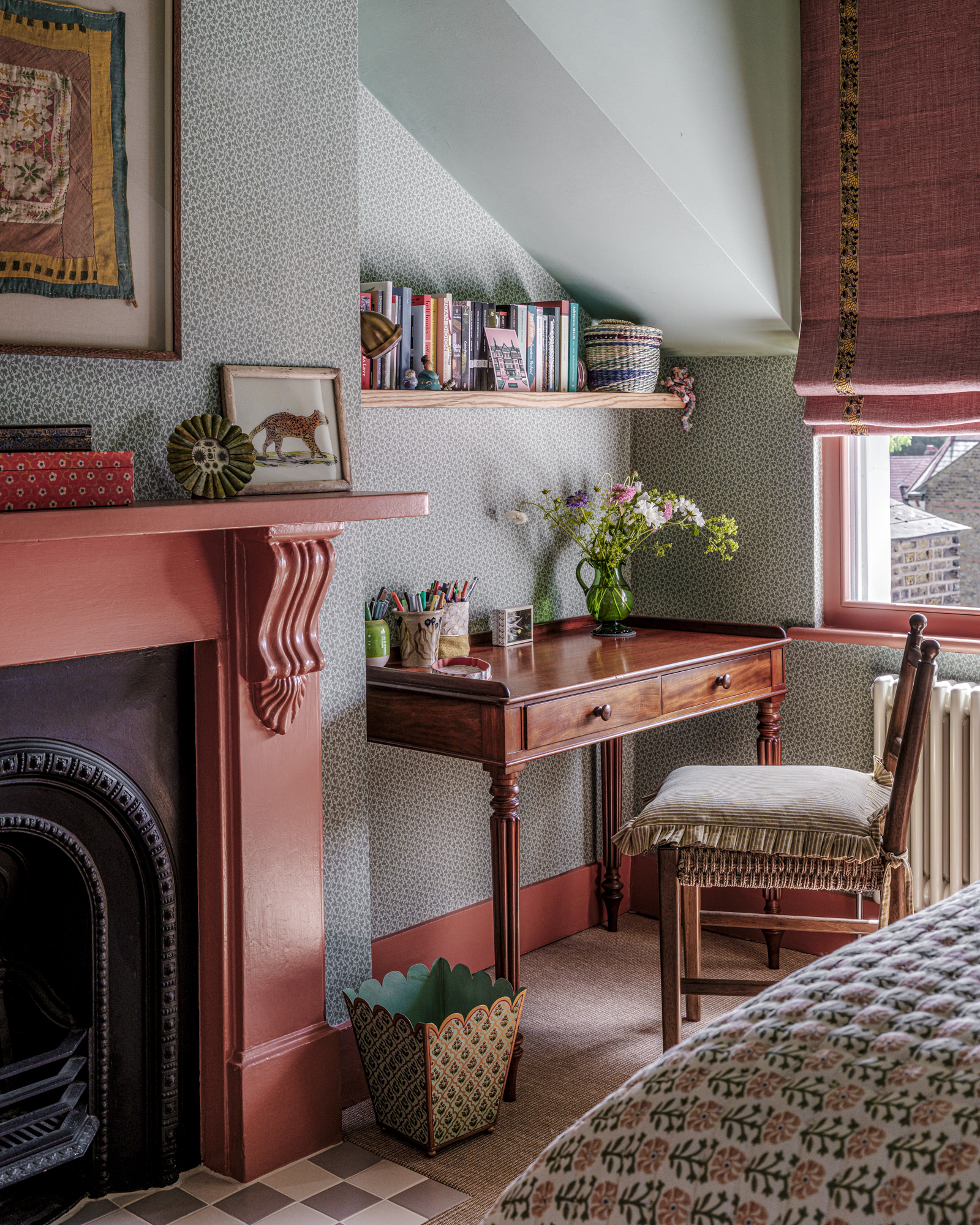 a bedroom corner with an antique desk and a wooden chair with a green vase with flowers on top and a wooden shelf above it with colourful books