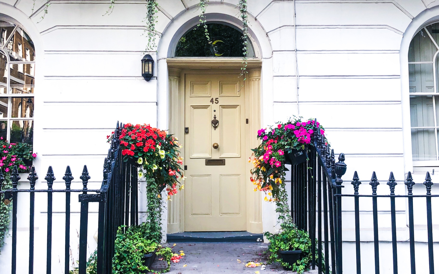 a white townhouse with a pale yellow front door, black iron gates with colourful flowers hanging from them