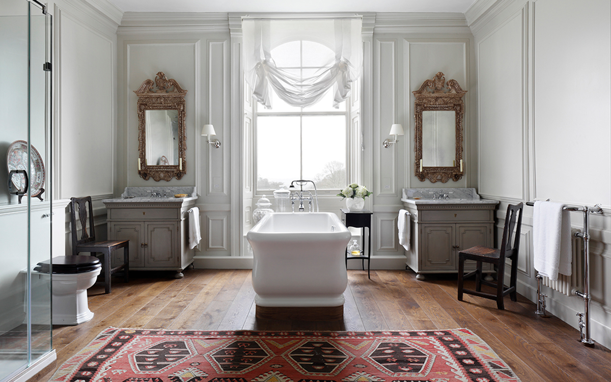a large grand bathroom with a white freestanding tub in the centre, a grey vanity unit in each corner behind the bath with ornate gold mirrors above and a red aztec patterned rug in the foreground