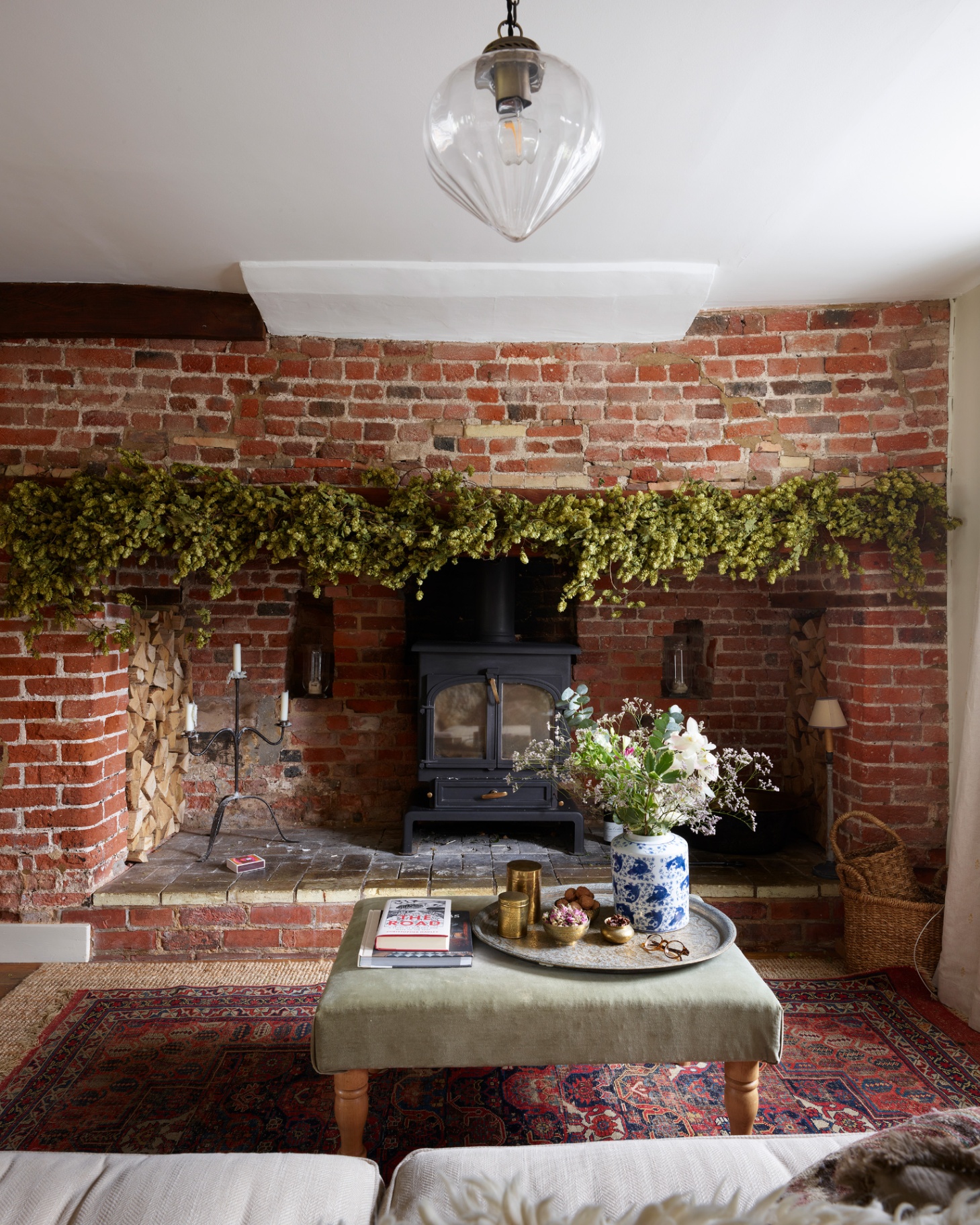 a living room with a large red brick fireplace with greenery hanging along it and a black wood burner inside and an upholstered coffee table infront of it