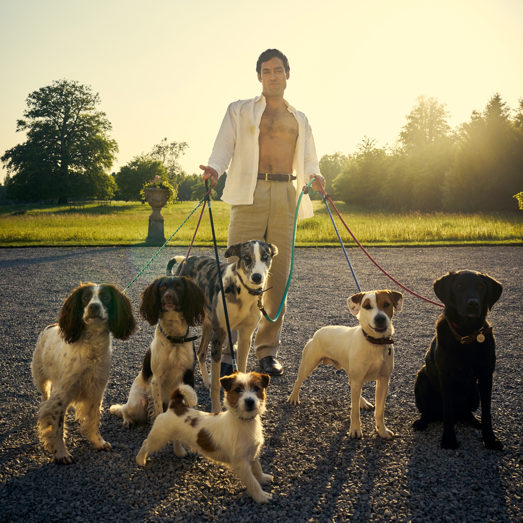 Alex Hassell as Rupert Campbell-Black standing with his white shirt open, holding 7 dogs of different breeds on leads