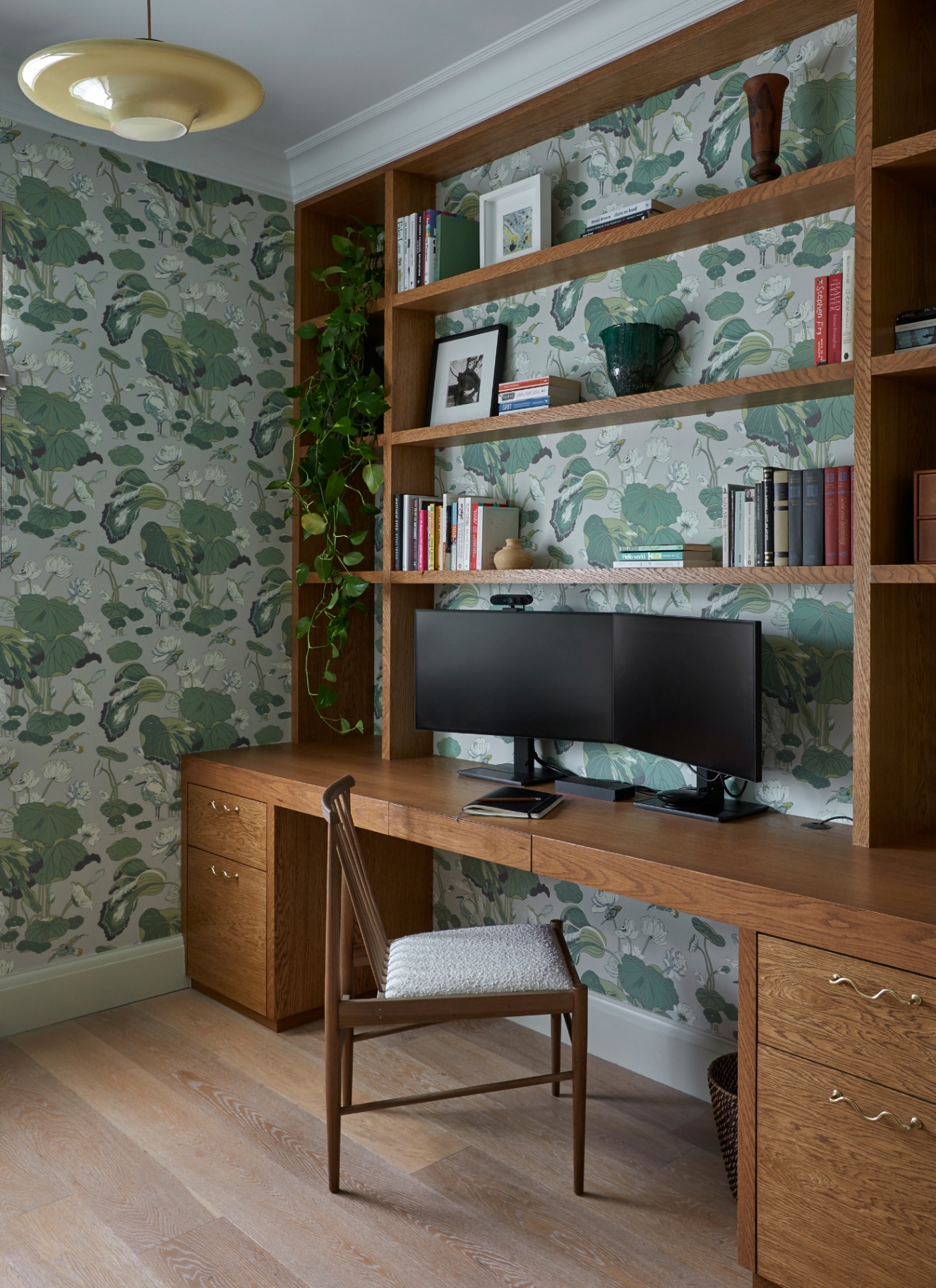 Custom oak built-in study unit framing dual computer monitors on a long wooden desk, with open shelving, books, and green botanical wallpaper background
