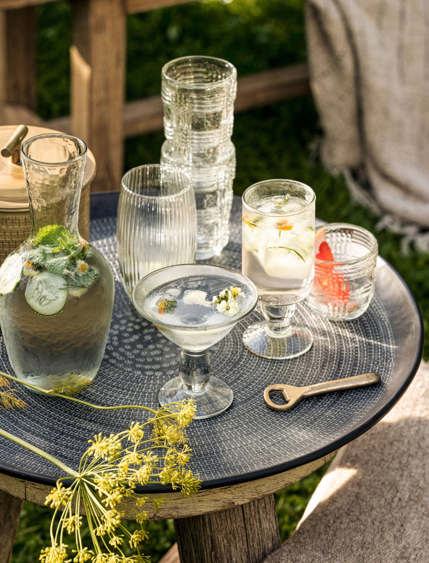 A wooden outdoor round table with glasses and a carafe of cucumber water in it