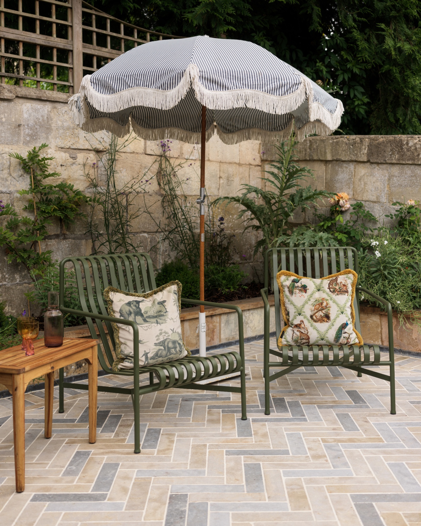 a patio with two dark green metal chairs, a cream patterned cushion on each chair with a white and blue fringed parasol between them