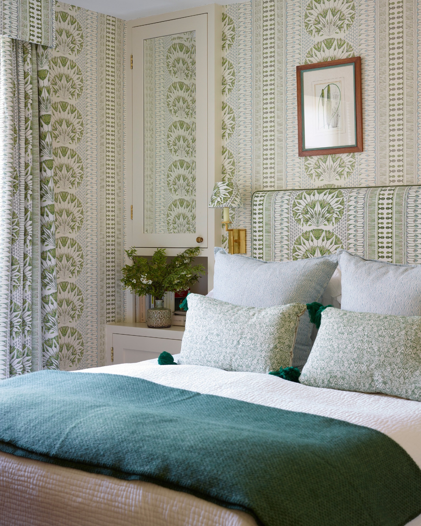 a bedroom with a green and white botanical pront wallpaper and matching sconce and headboard, a white built in mirrored cupboard next to the bed and a dark green throw at the end of the bed