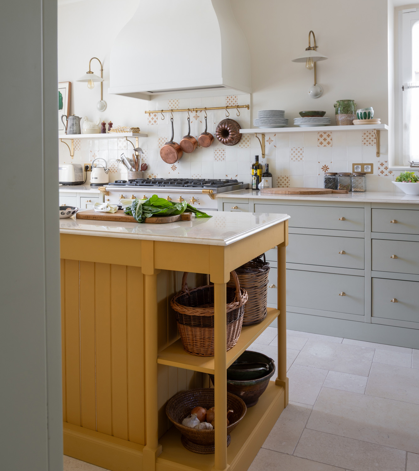 a kitchen with white walls with shelves and copper pans hanging, pale sage cabinetry and a mustard yellow kitchen island with shelves underneath