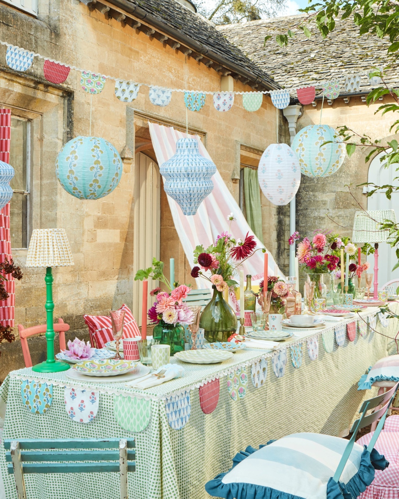 an outdoor dining table with a green polka dot tablecloth, metal dining chairs with seat pads, patterned bunting and paper lanterns hanging above