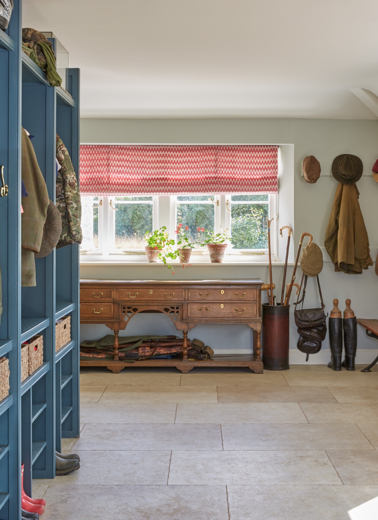 a boot room with a blue open cupboard, an antique table with storage, an umbrella storage holder and a large window with a white and pink blind