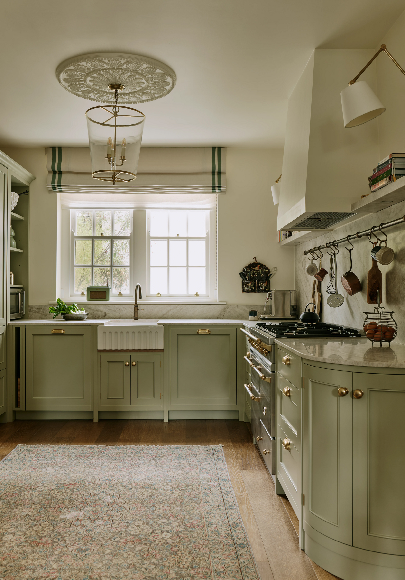 A kitchen with sage green cabinetry, ribbed butler sink and range cooker, with a grey marble backsplash. A floral rug is situated in the middle of the floor and above is a pendant lantern 