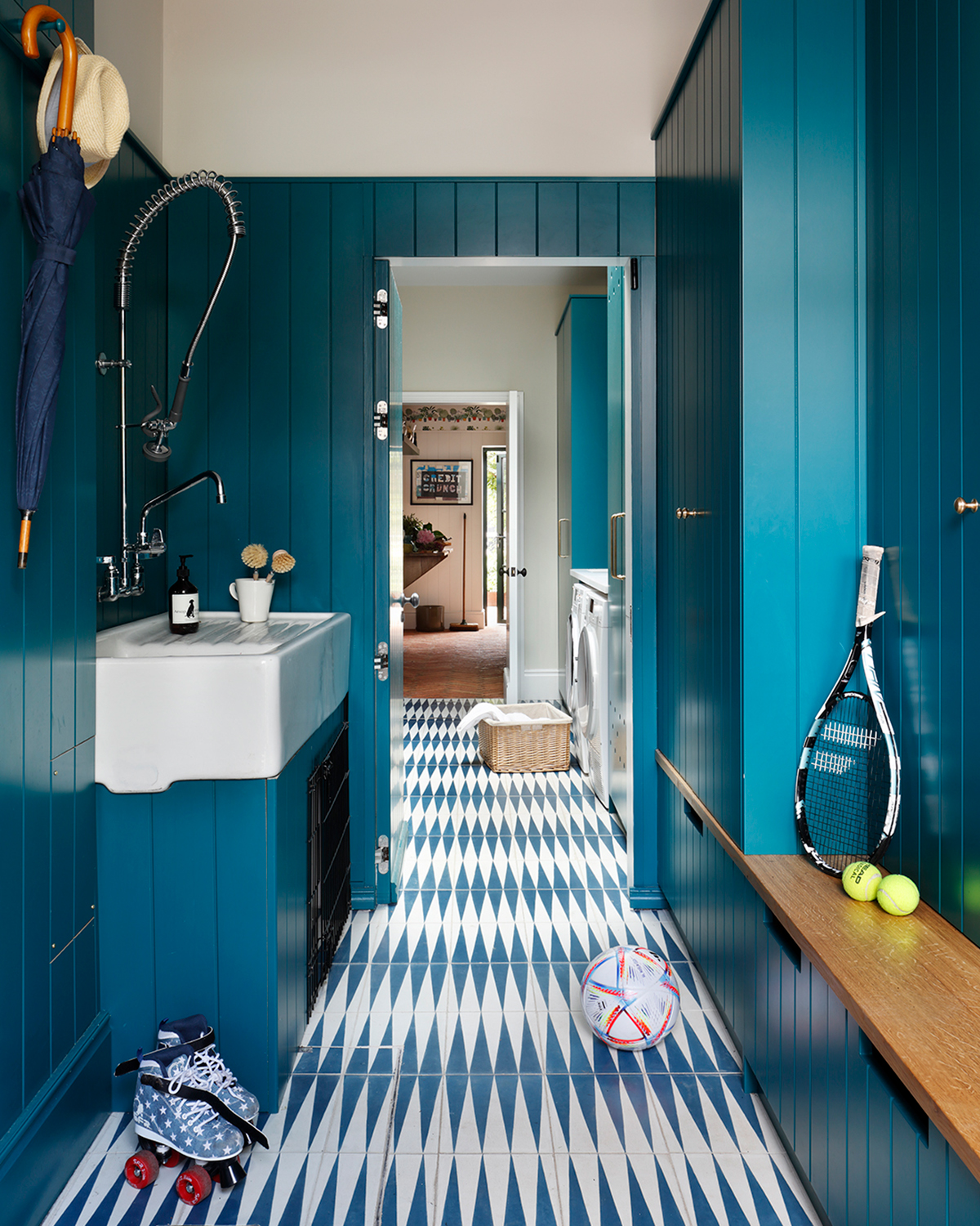 a boot room with bright teal tongue and groove walls, a teal and white harlequin pattern floor, a large sink with a silver shower above it, a pair of roller skates on the floor and a tennis racket and balls on a wooden bench