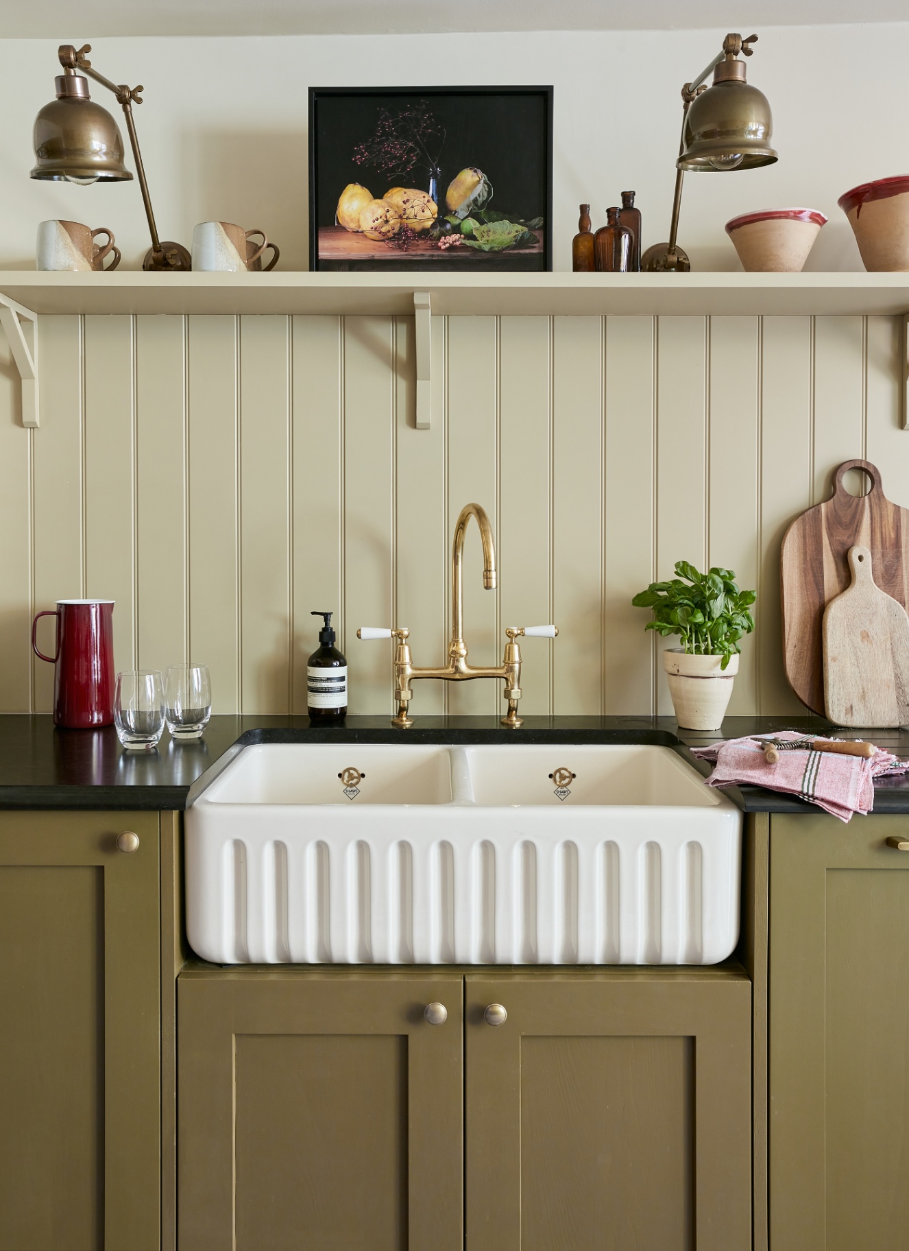 a kitchen sink area with olive green painted cabinets, cream tongue and groove panelled walls, a belfast sink with gold hardware and a shelf with two brass lams and a painting