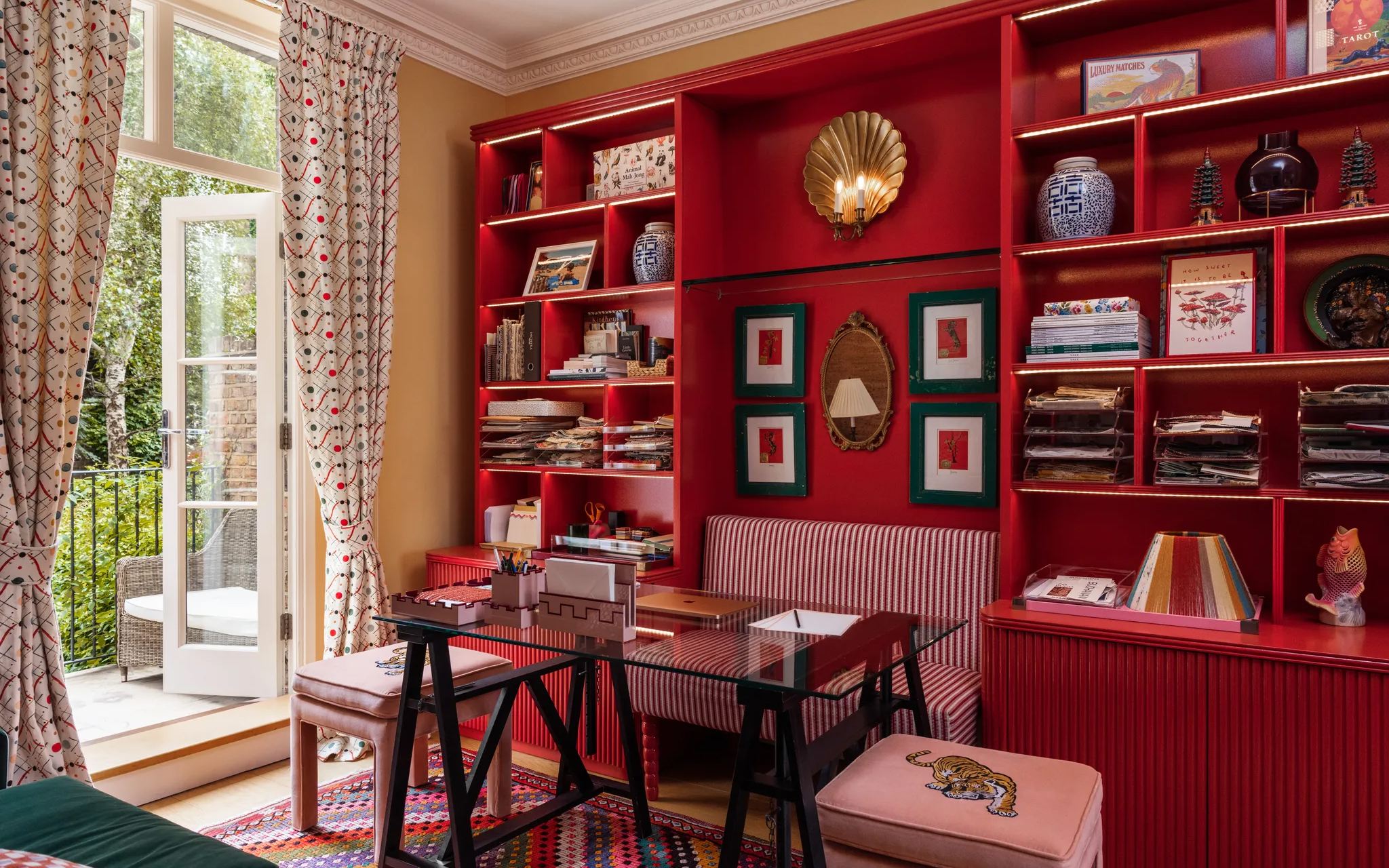 Home office featuring bright red built-in shelving with integrated lighting, a glass-topped desk, pink velvet footstools with embroidered tiger details, red-and-white striped banquet seating, and patterned curtains by a French door opening to a garden