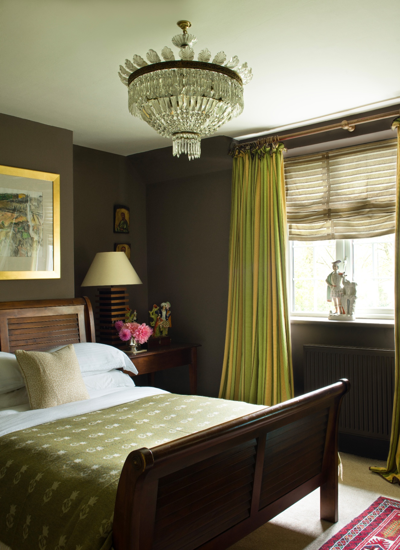Moody dark chocolate brown bedroom with olive green curtains, wooden sleigh bed, patterned green throw, crystal chandelier, and a bedside lamp on a wooden table