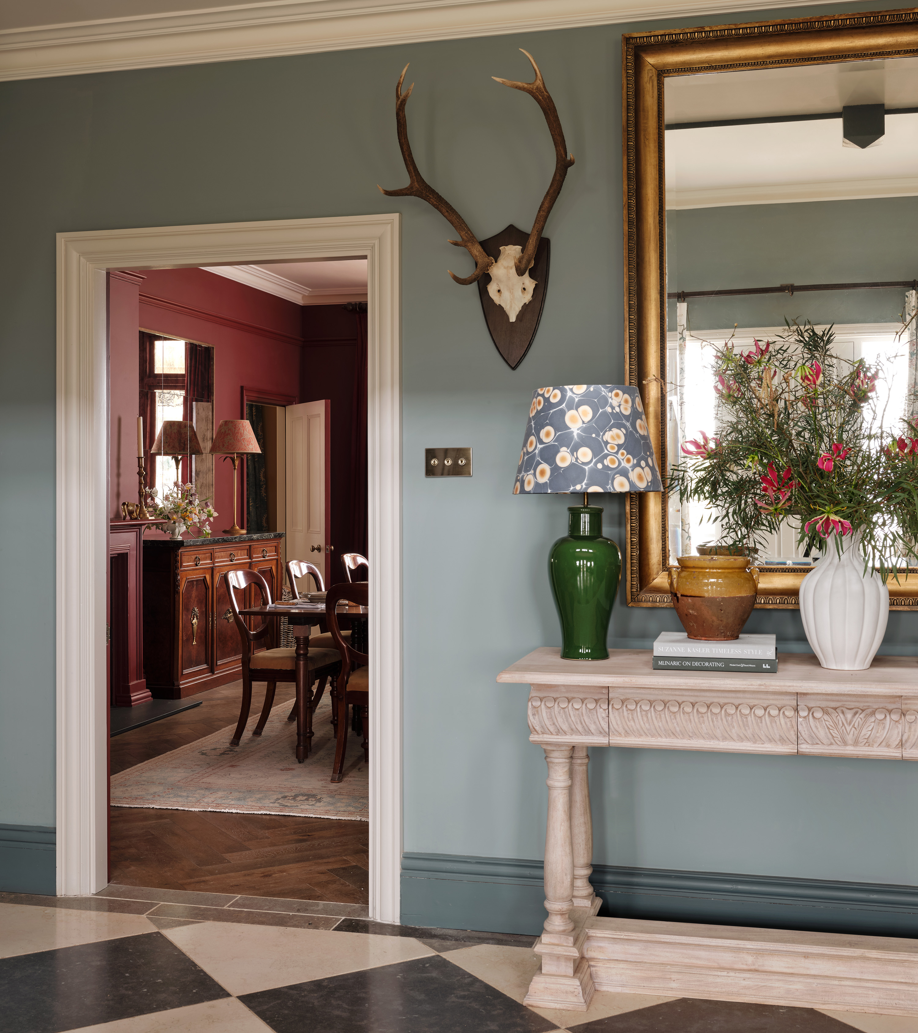 a hallway with pale blue walls, black and white checkered floor and a stag skull on the wall looking through to a dark red dining room