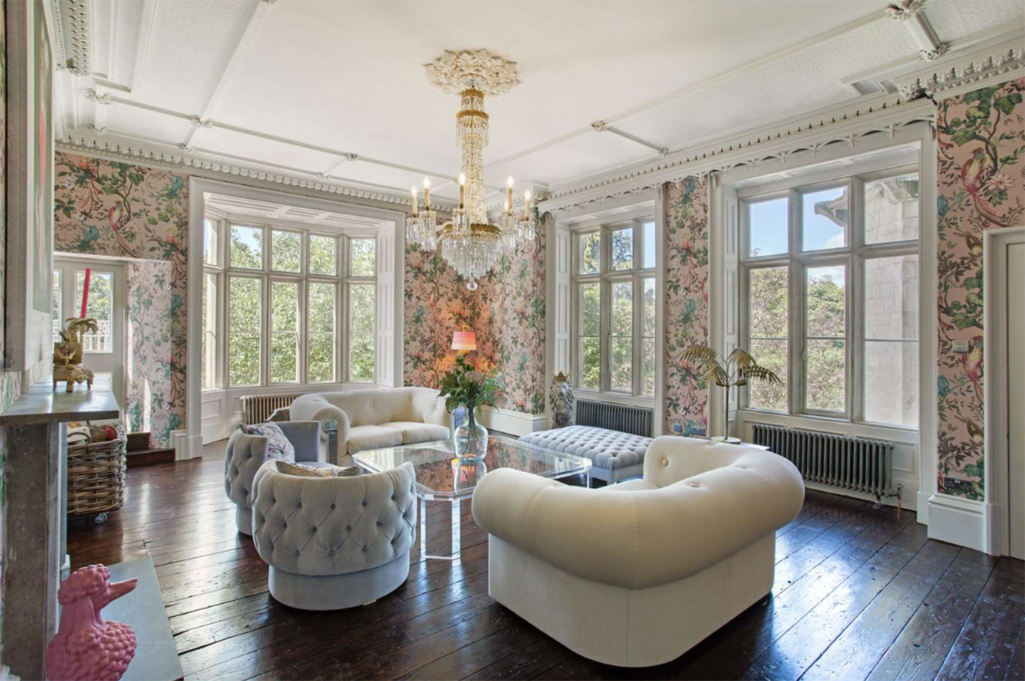 a living room with pink floral wallpaper, cream and grey sofas and a large crystal chandelier