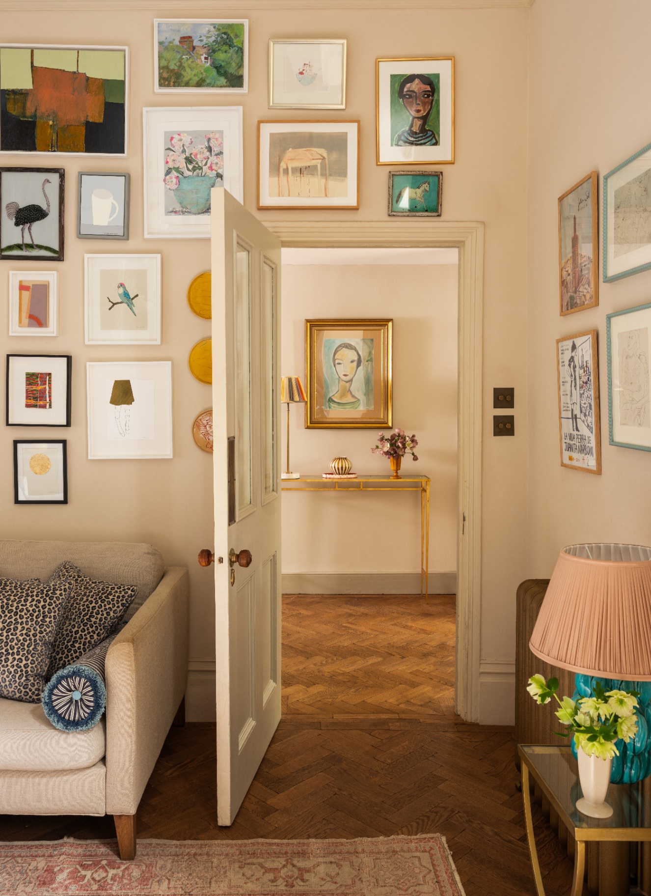 a living room corner with a gallery wall, wooden parquet flooring and an open door looking onto the hallway with a framed painting above a console table