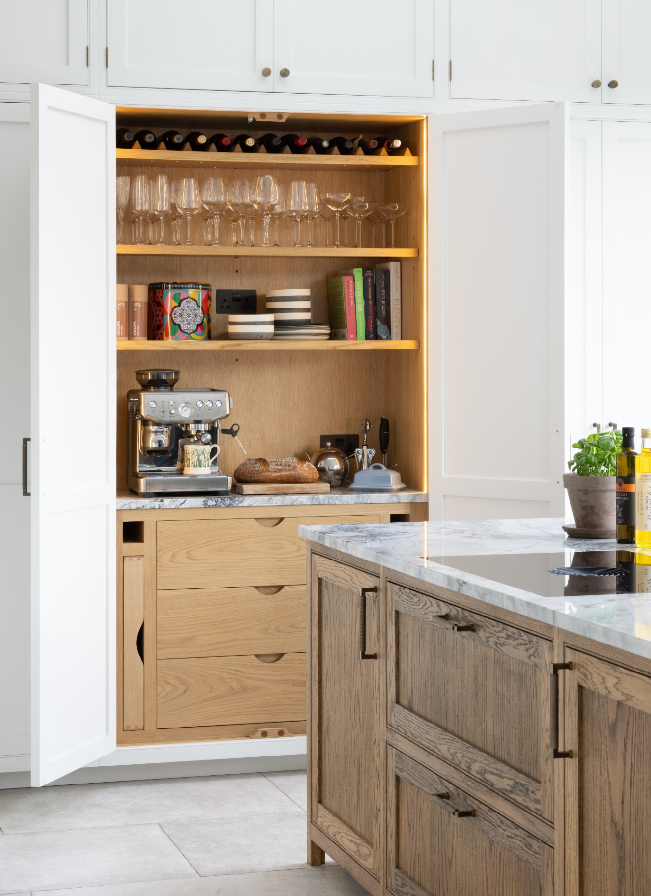 Full-height white pantry cabinet with open bi-fold pocket doors revealing a warm oak interior, backlit glassware shelves, wine rack, espresso machine, and wooden drawers