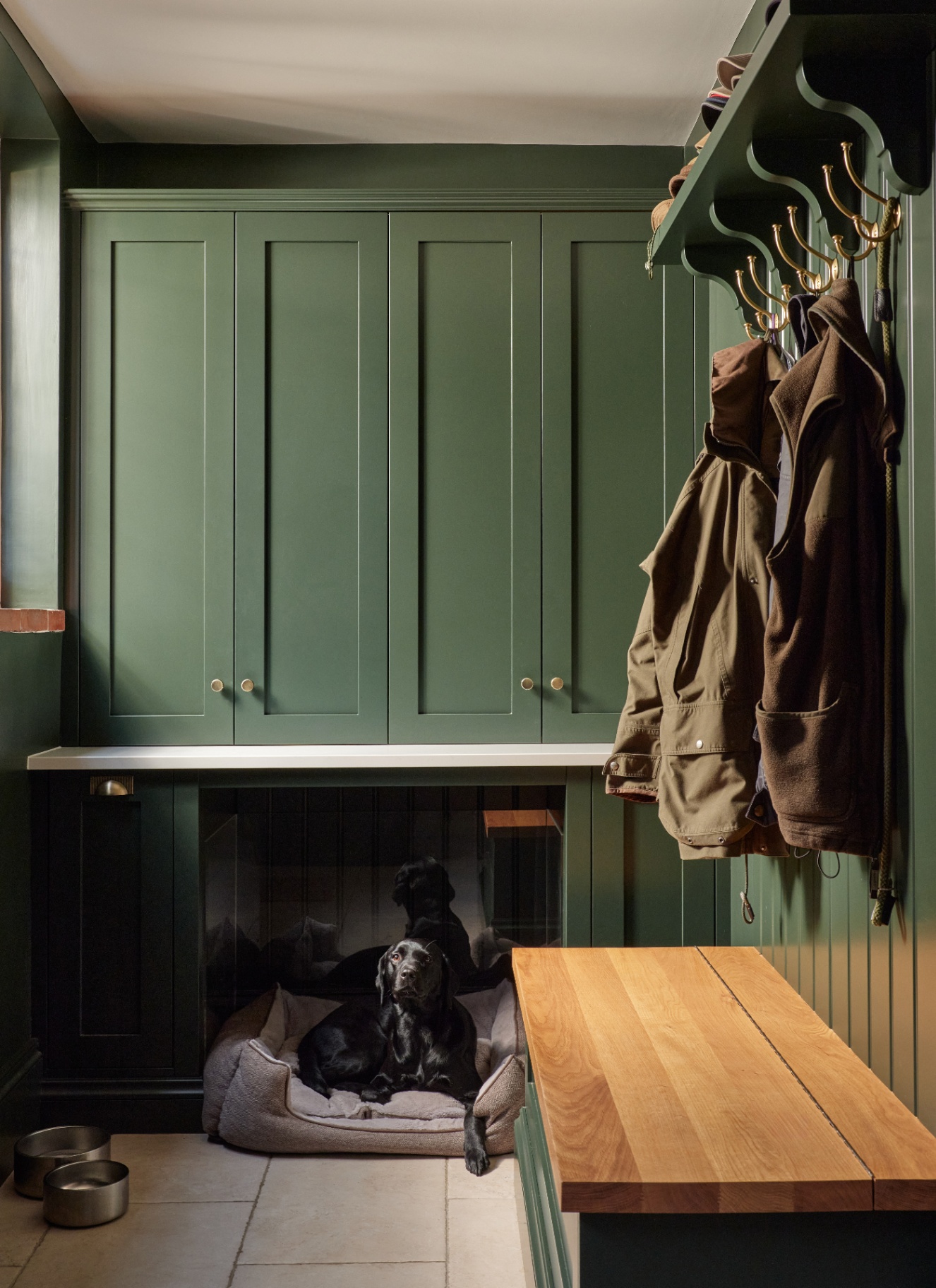 a boot room with dark green cabinetry, a dog bed nook with a black labrador inside and a hanging rail with coats