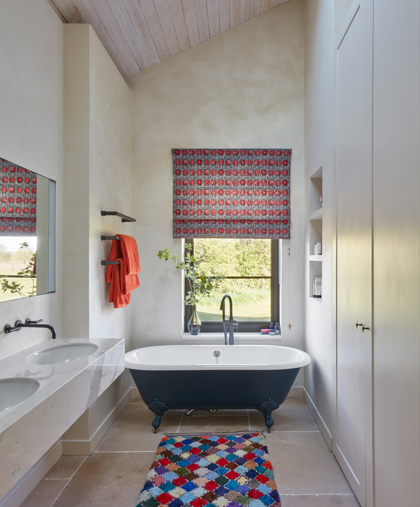 A high-ceilinged bathroom with a floating marble double basin on the left, with a mirror above, and built-in cabinets on the right. A freestanding black tub sits under a window at the end of the room, and a colourful rug tops the large floor tiles