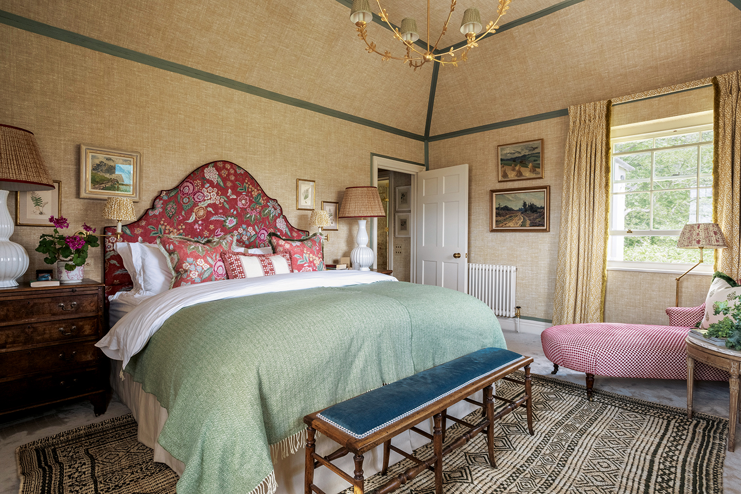 A large bedroom with hemp vinyl wallpaper with dark blue borders on the corners of the room and angles of the sloped ceiling. A double bed with a floral red headboard sits on a patterned monochrome jute rug with a bench at the end and wooden bedside chests