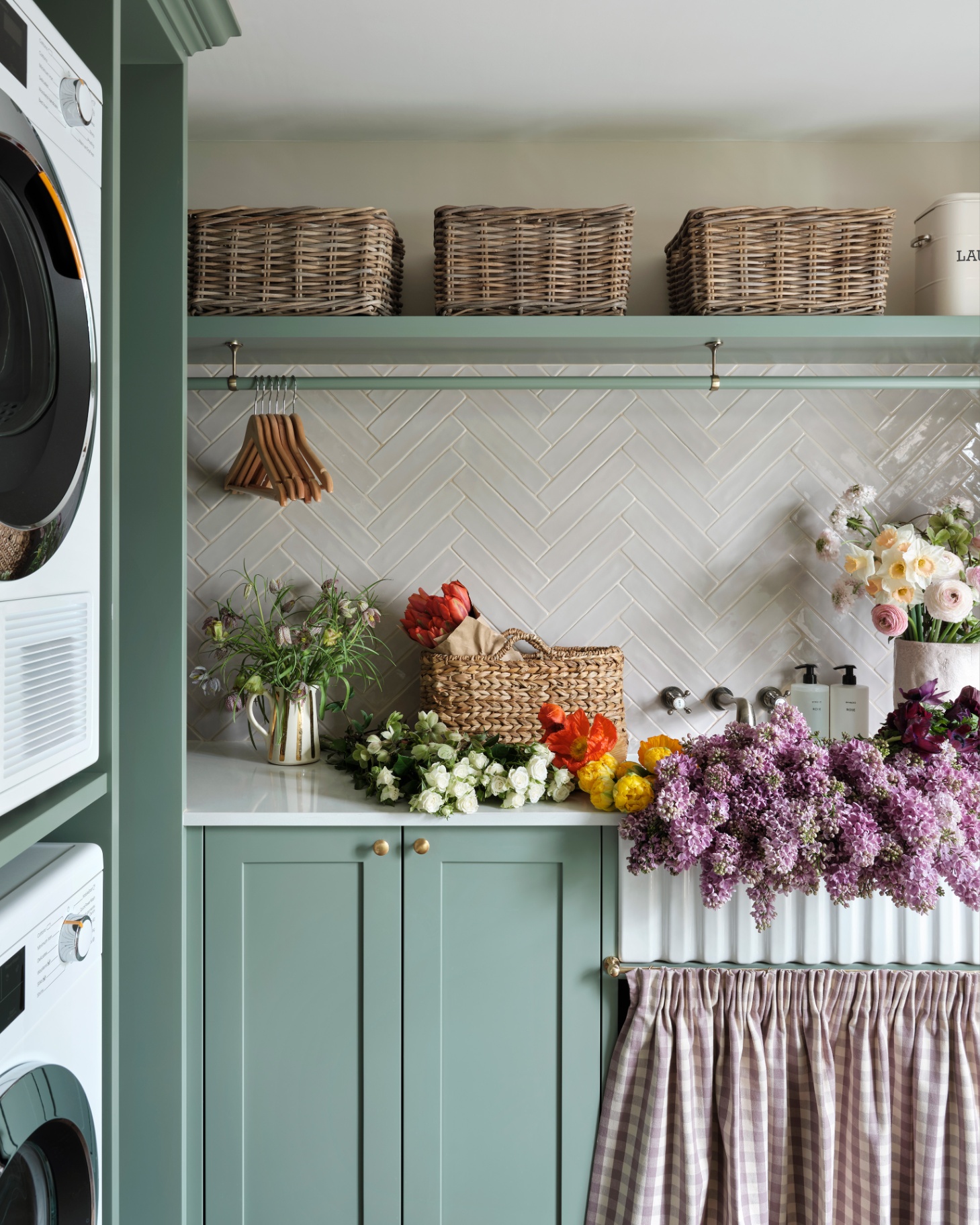 a utility room with stacked washing machines, sage green cabinetry, a high open shelf with wickers baskets and lots of colourful cut flowers on the worktops