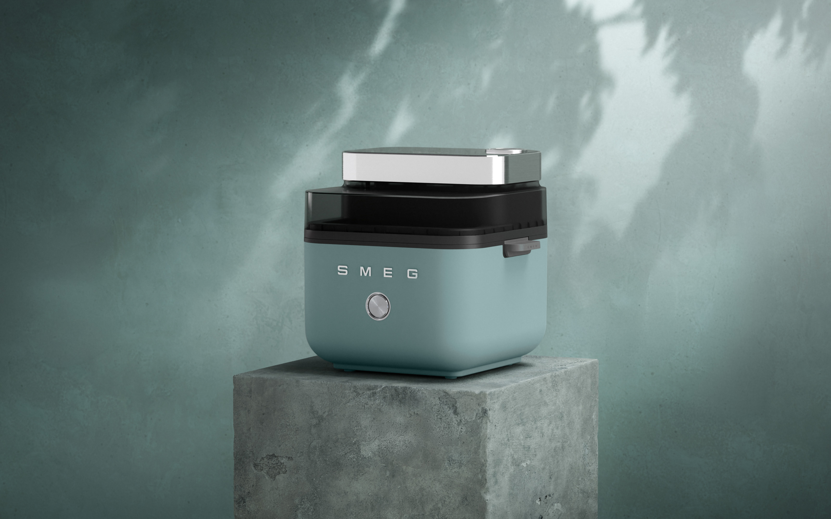 Matte sage green SMEG countertop air fryer displayed on a concrete block. The retro-style appliance features silver controls, a transparent glass middle section, and a silver top lid, set against a atmospheric green wall with soft shadows