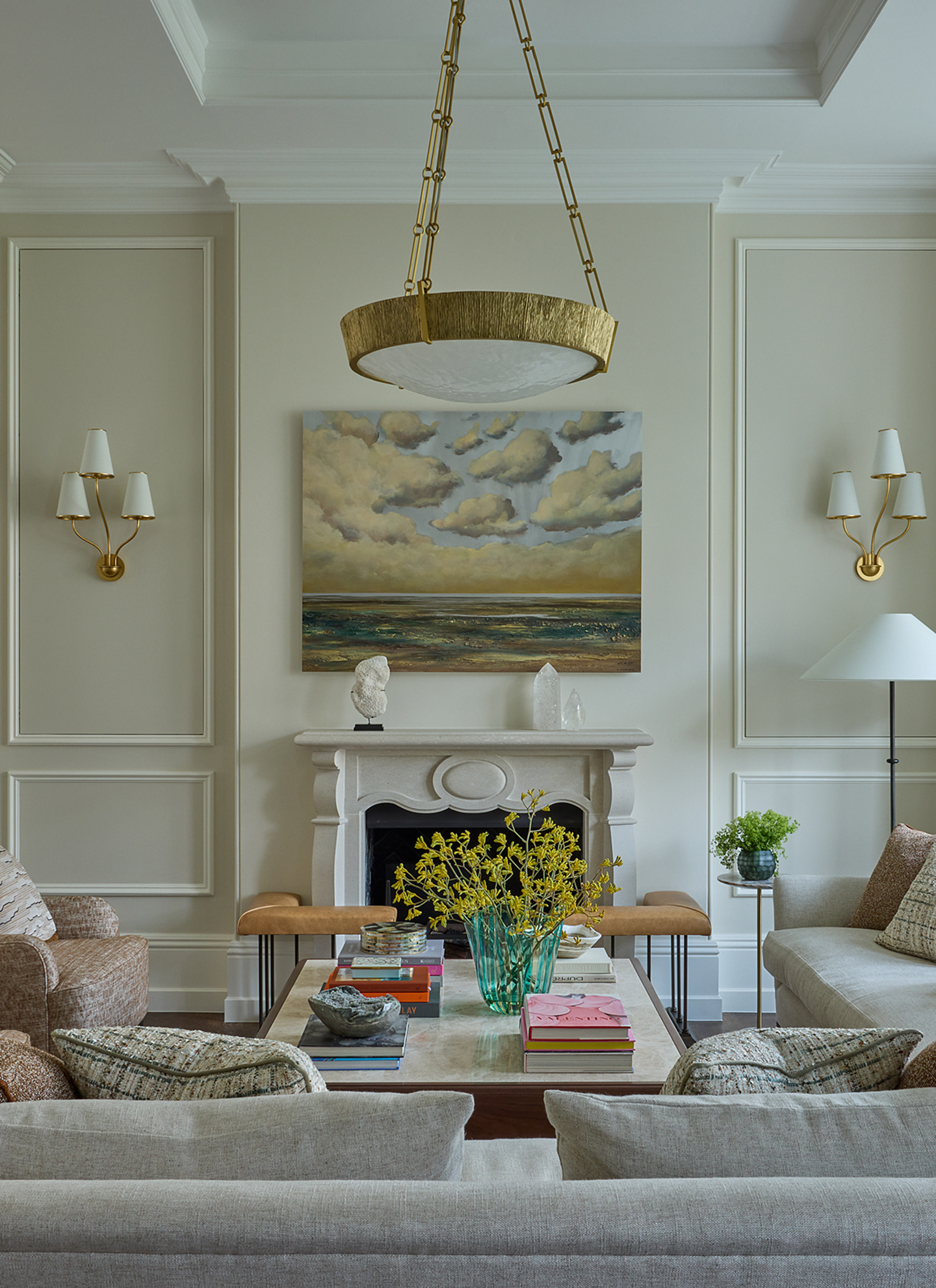 An elegant neutral drawing room featuring a stone fireplace framed by gold wall sconces, a large brushed-brass pendant light suspended above a coffee table, and soft upholstered sofas in warm beige tones.